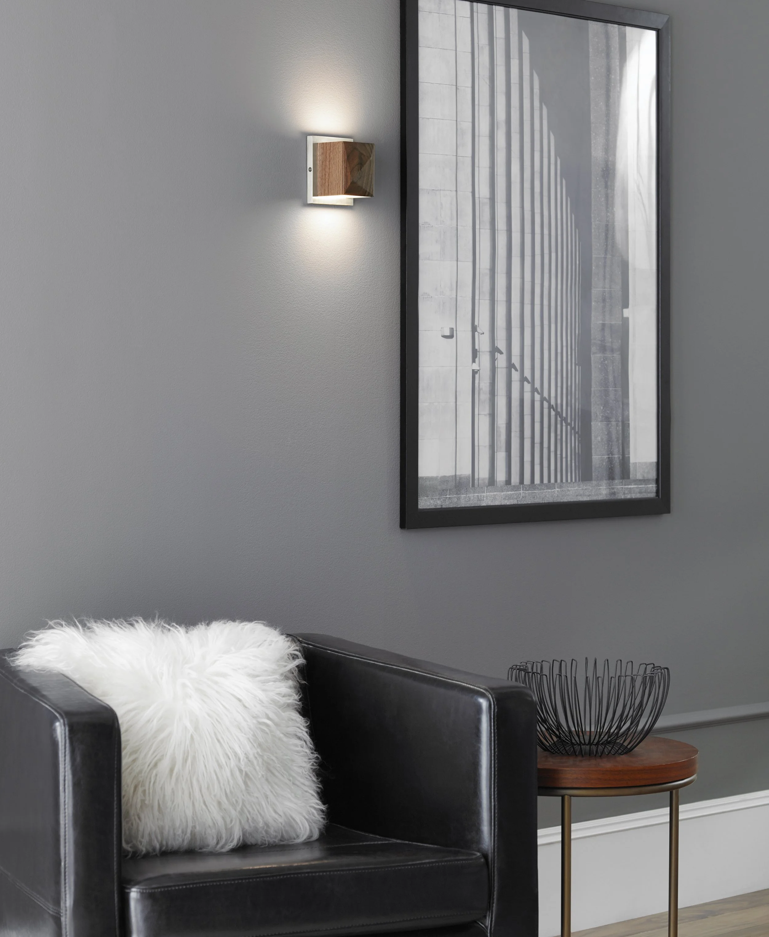 Modern living room with a black leather armchair, white furry pillow, wall sconce, framed architectural artwork, and round side table with a black wire basket.
