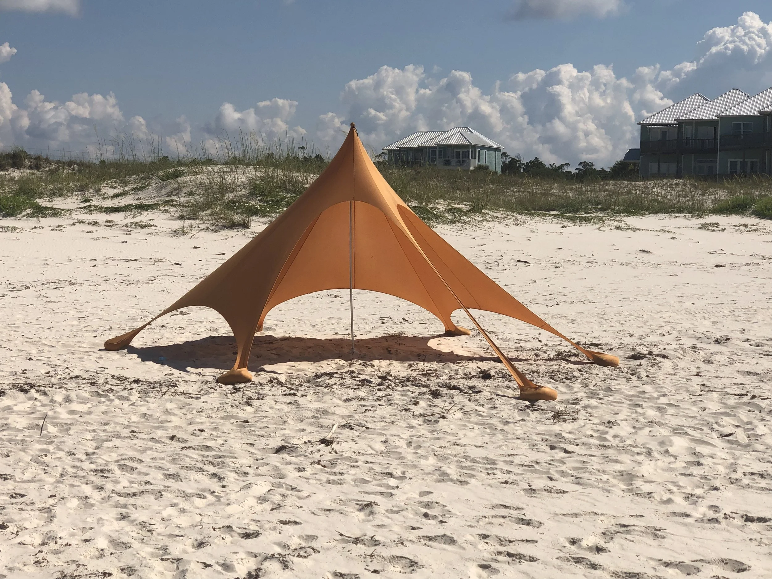Orange shade canopy on a white sandy beach with coastal houses, dunes, and cumulus clouds.