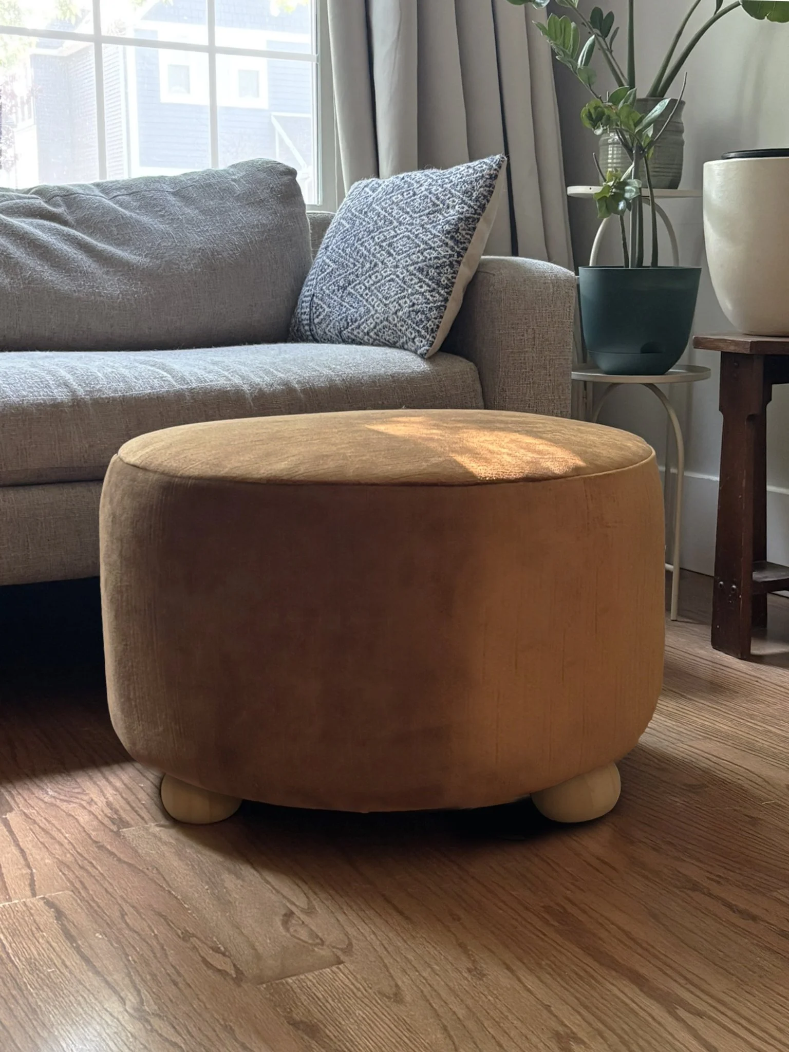 Round brown upholstered ottoman in front of a gray sofa with patterned pillow and potted houseplants