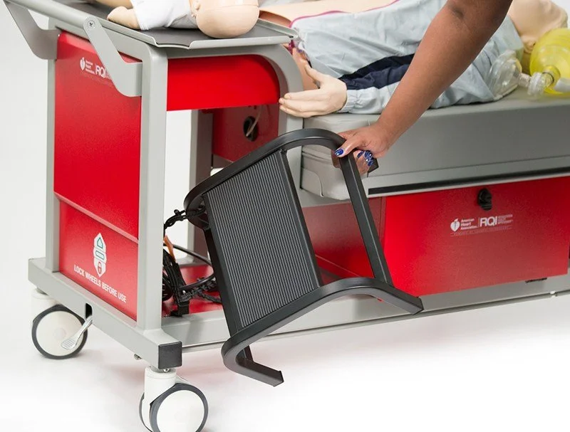 Healthcare worker attaching a black support accessory to a red medical training cart with a patient simulator mannequin