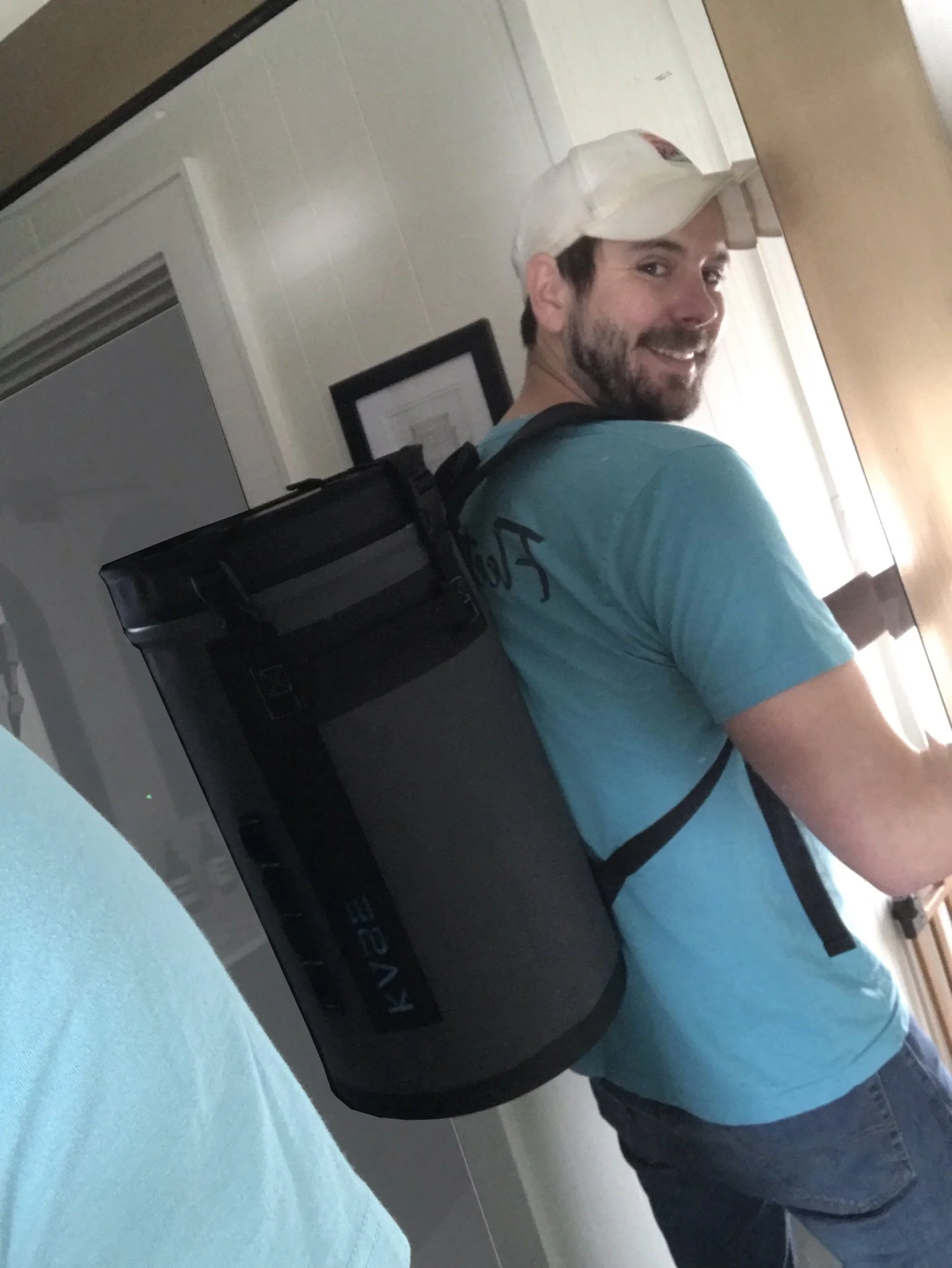 Smiling man wearing a white baseball cap and blue shirt while carrying a large black delivery bag indoors