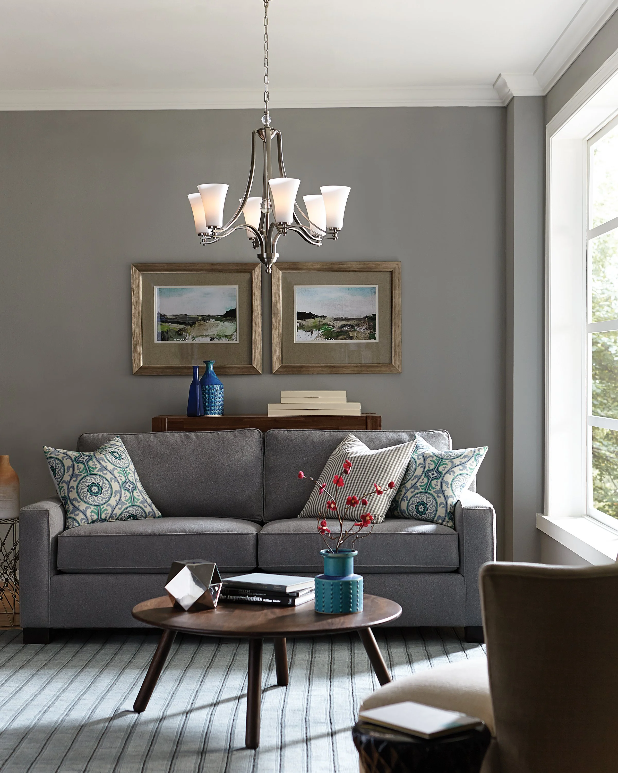 Modern gray living room with a chrome chandelier, gray sofa and patterned pillows, framed artwork, coffee table, and large windows.