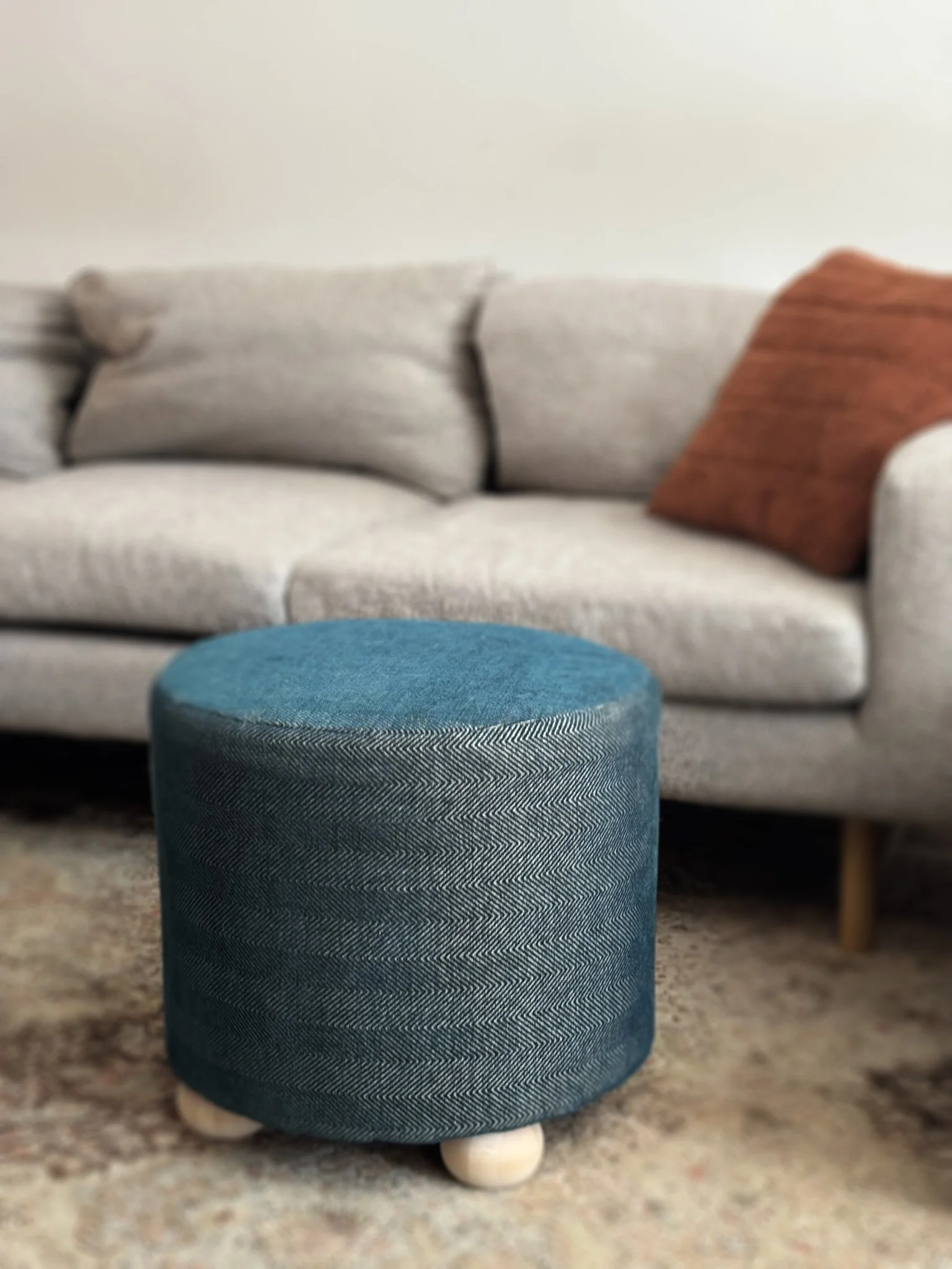 Blue cylindrical upholstered ottoman with a chevron pattern and wooden legs in front of a beige sofa with pillows