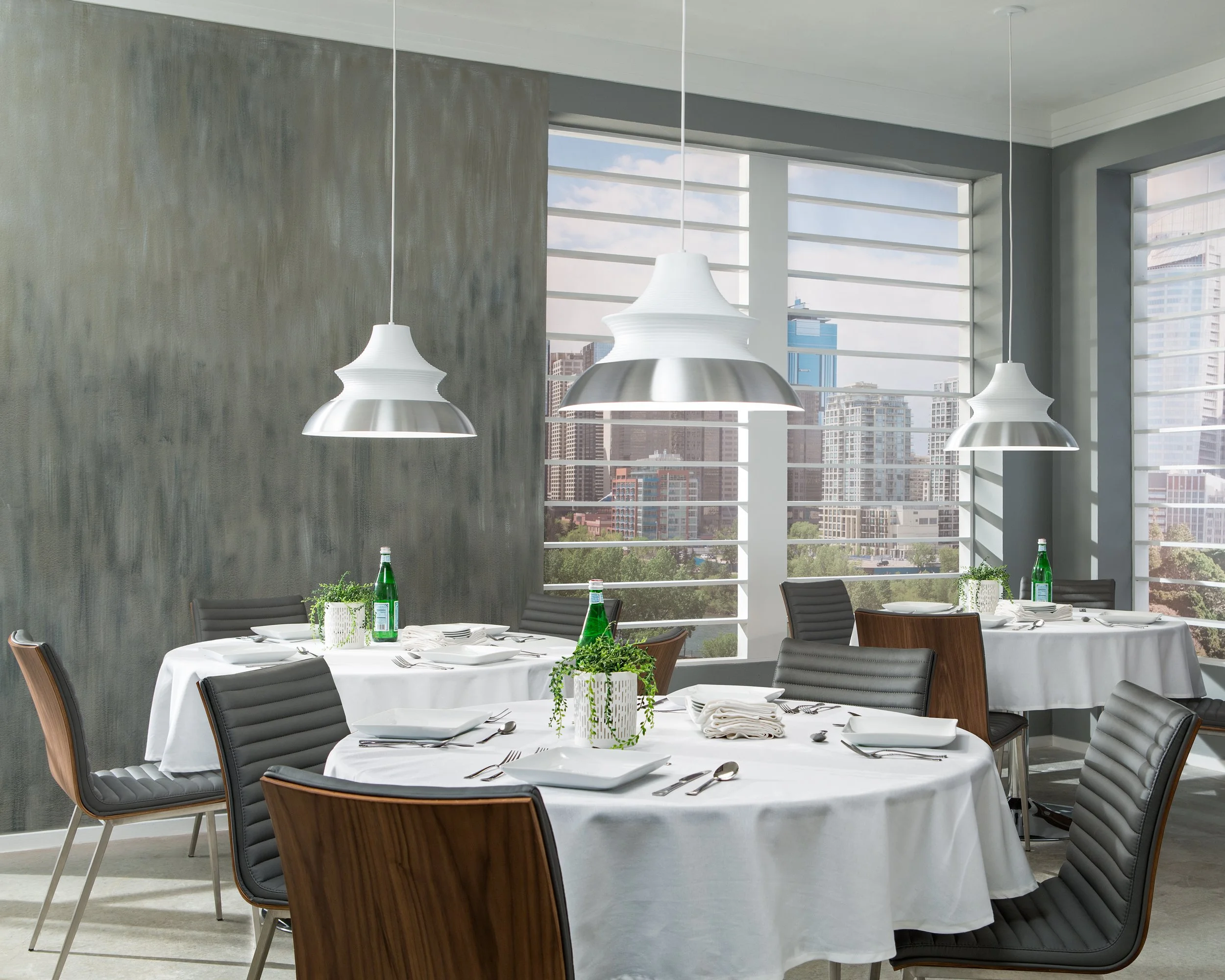 Modern restaurant dining room with white tablecloths, pendant lights, green plants, and city views through large windows