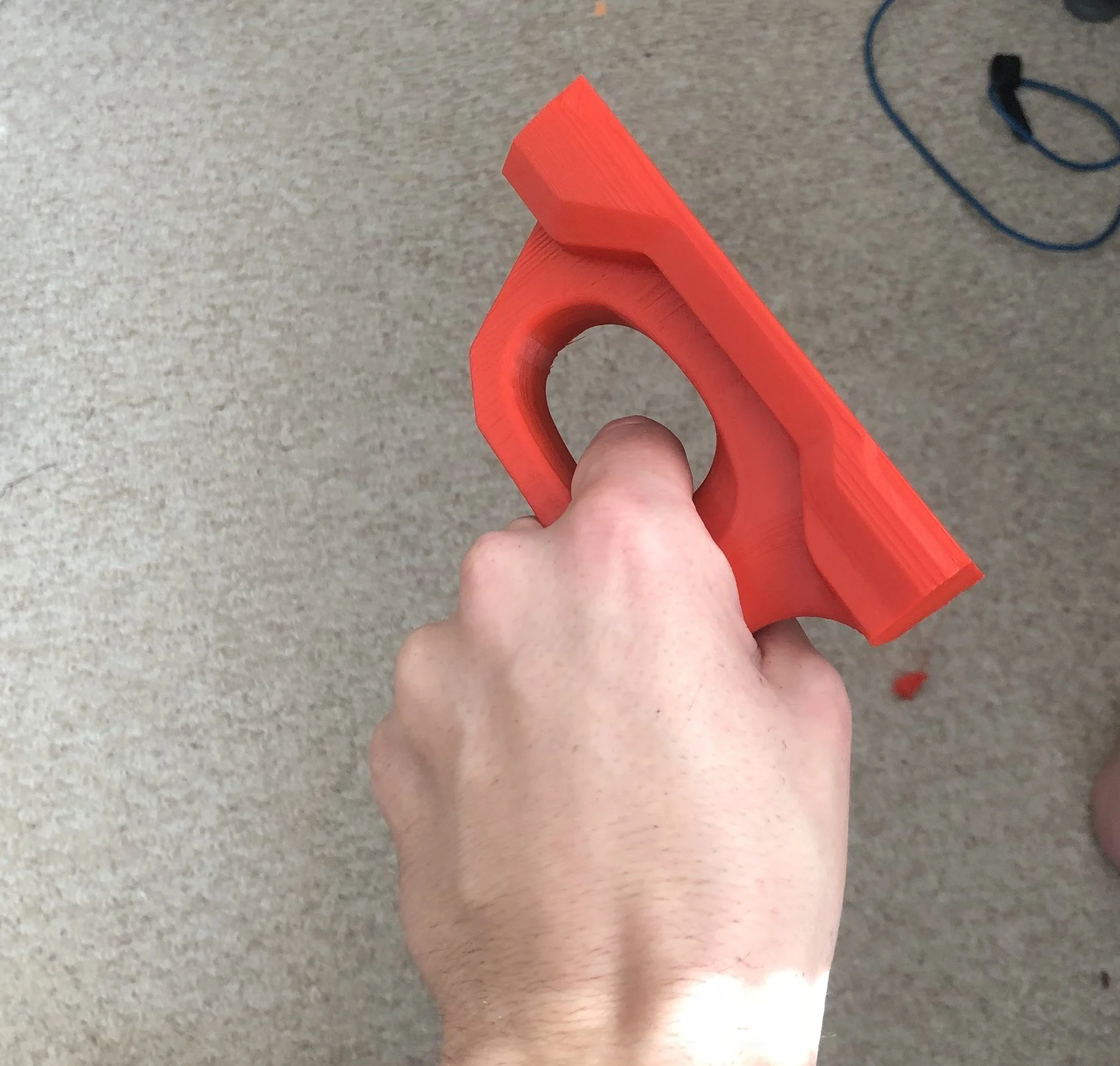 Hand holding a red 3D-printed plastic bracket over a carpeted floor