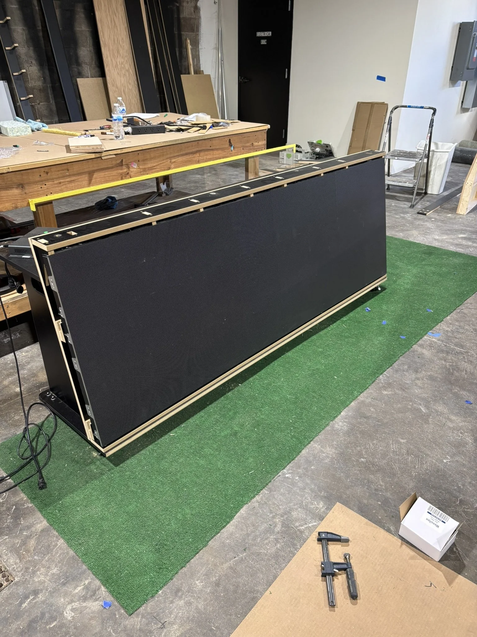 Large black electronic display panel being assembled in a workshop with tools, lumber, and a workbench