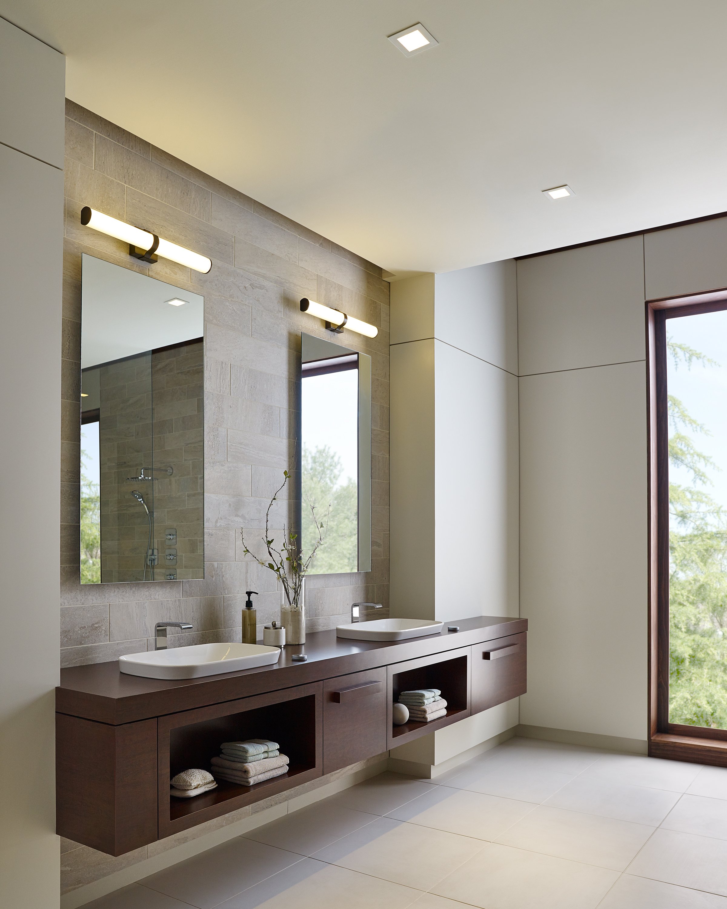 Modern bathroom with double vessel sinks, floating wood vanity, large mirrors, stone tile backsplash, and a sunlit window.