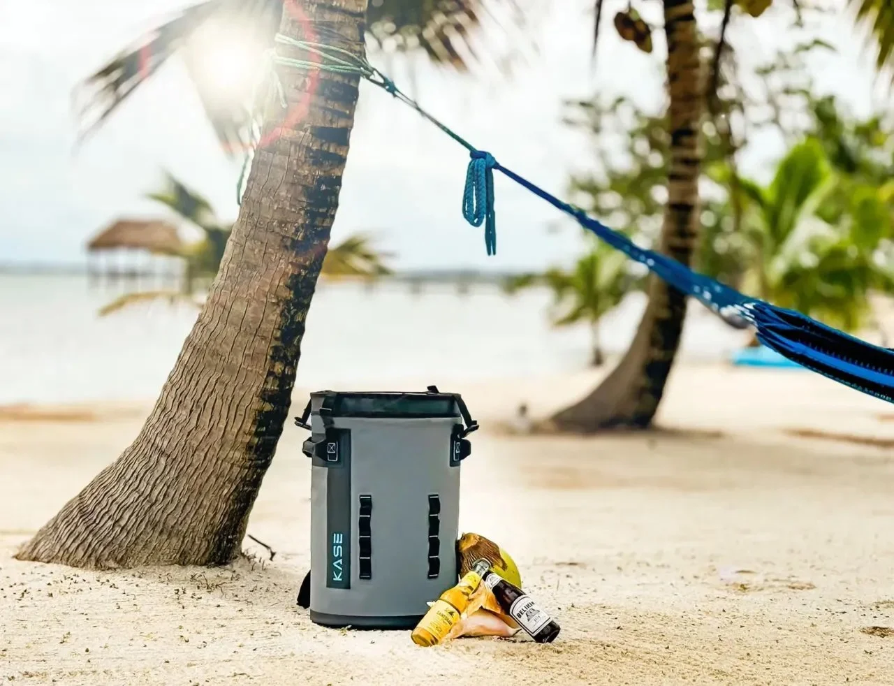 Portable cooler, beer bottles, and blue hammock on a tropical beach beneath palm trees