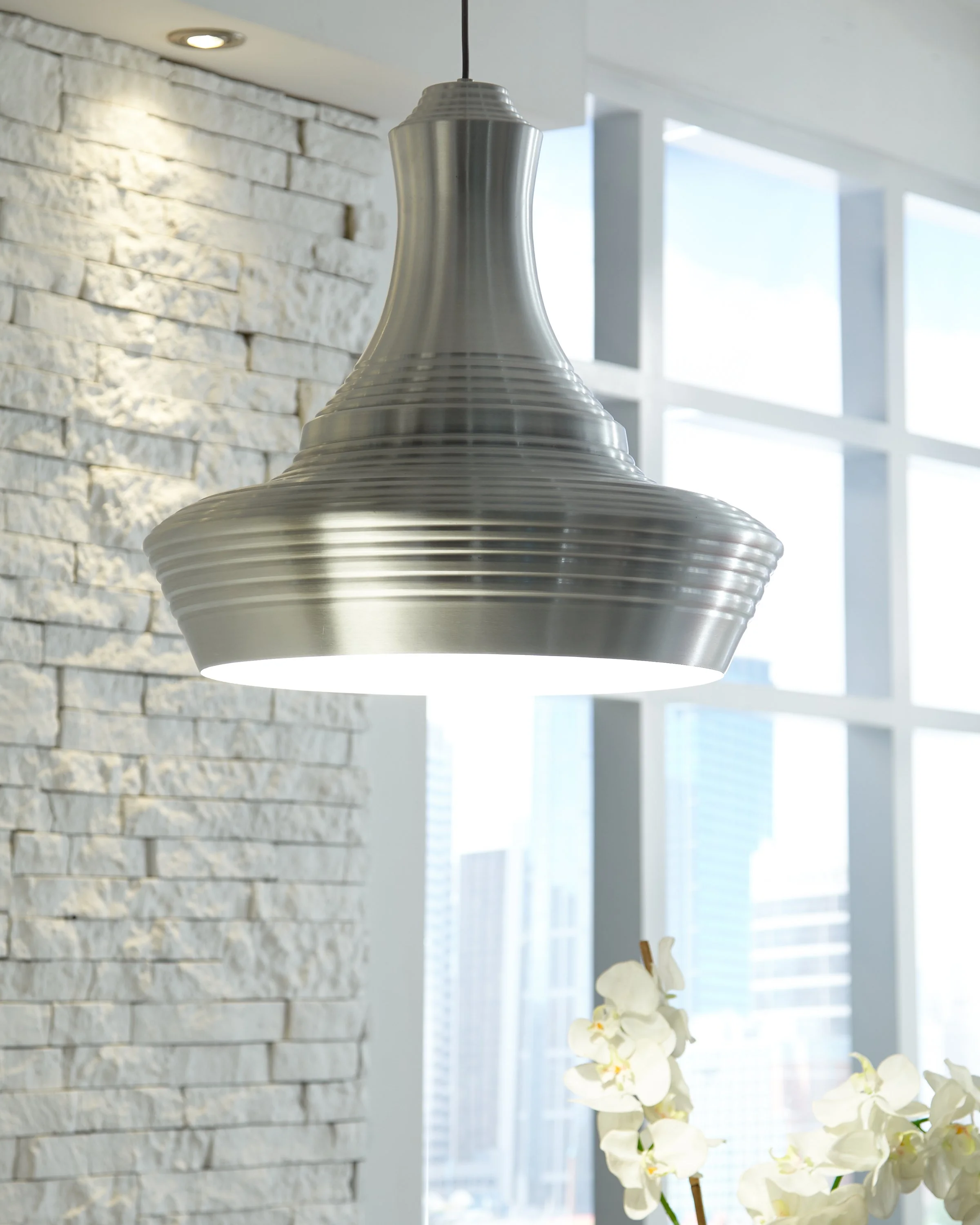 Modern brushed-metal pendant light hanging in a bright interior with a white brick wall, large windows, and orchids.