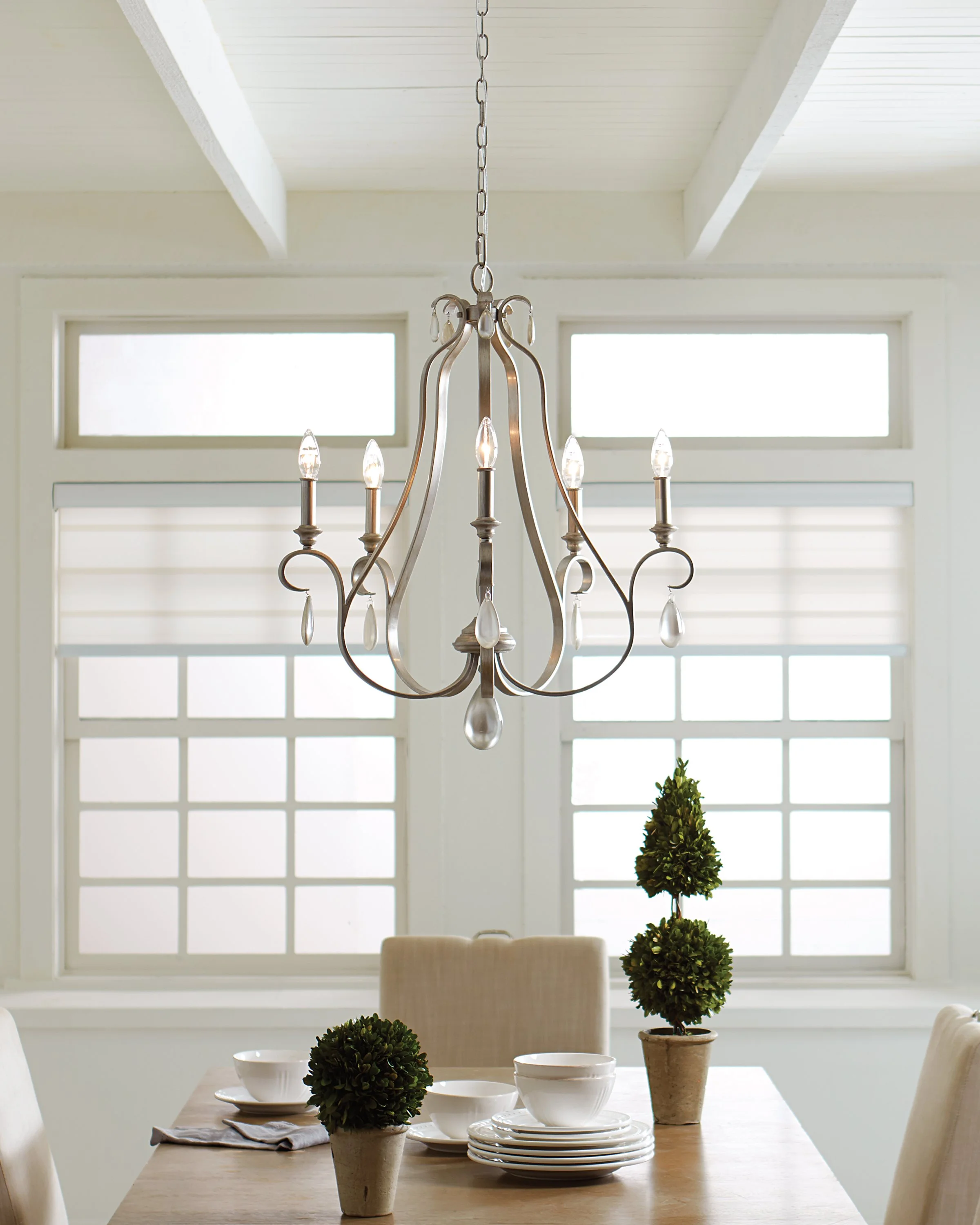 Elegant dining room with a crystal chandelier, white-paned windows, wooden table, white dishware, and potted topiary plants.