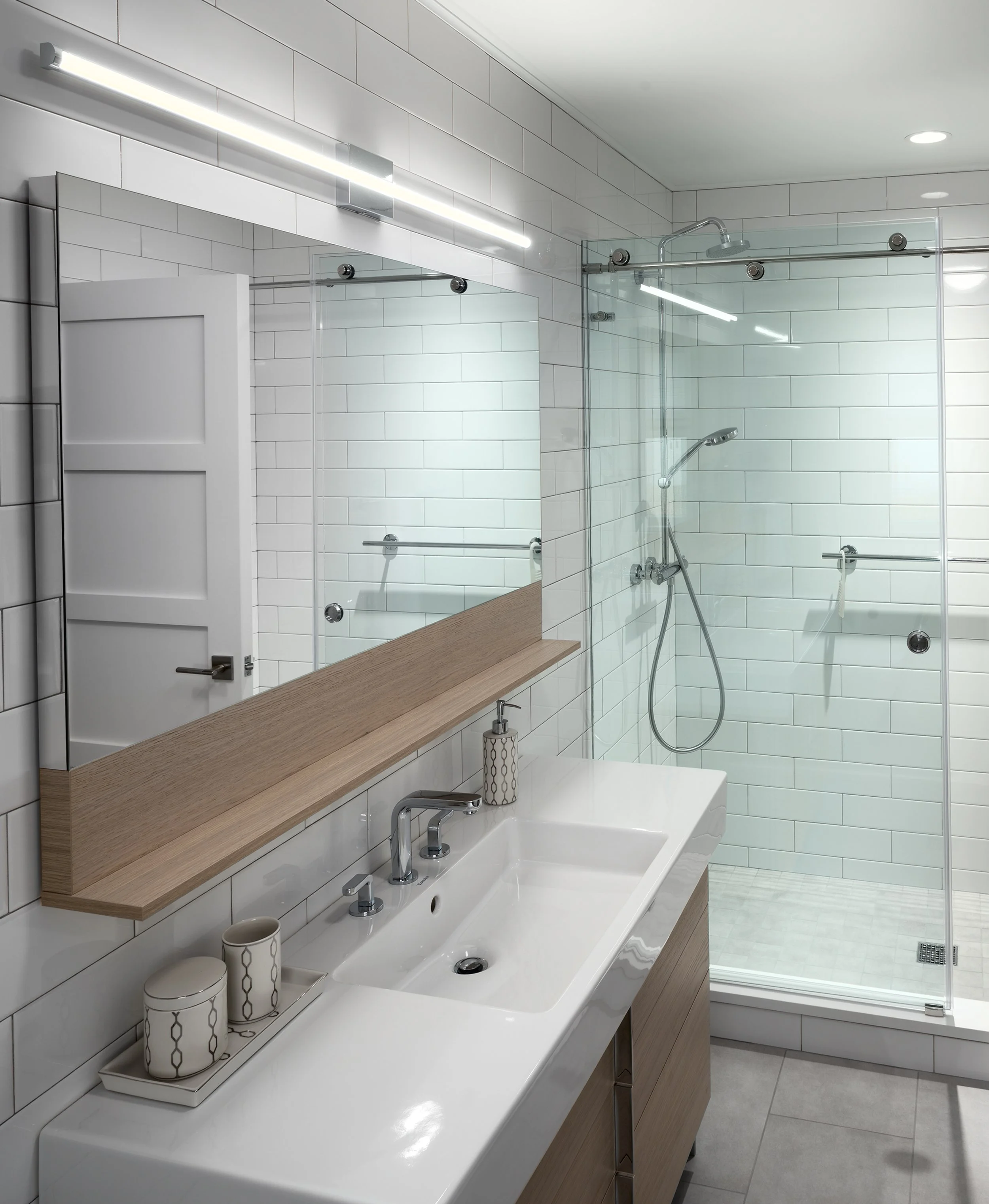 Modern white-tiled bathroom with a double sink vanity, large mirror, and glass-enclosed walk-in shower with rainfall showerhead.