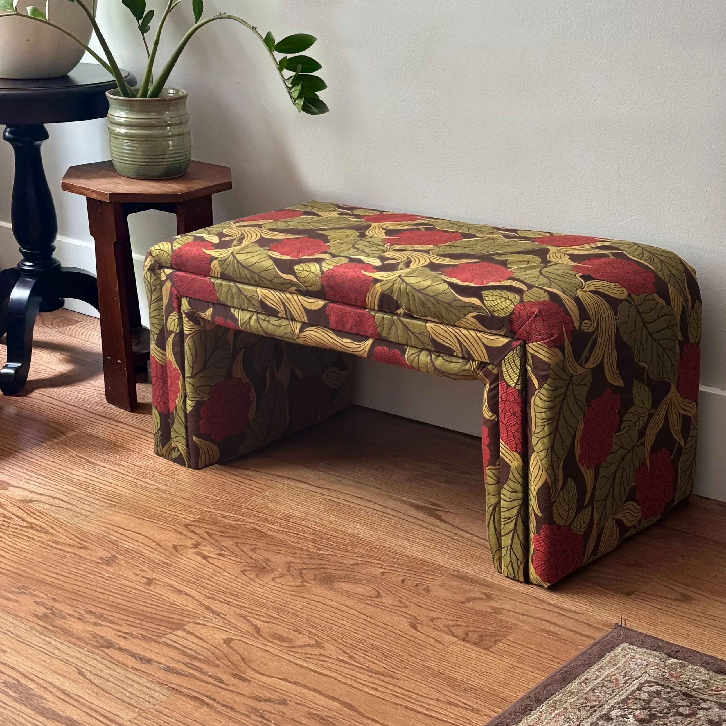 Small wooden table draped with a red, olive, and brown floral tablecloth beside a potted green plant and black pedestal table.