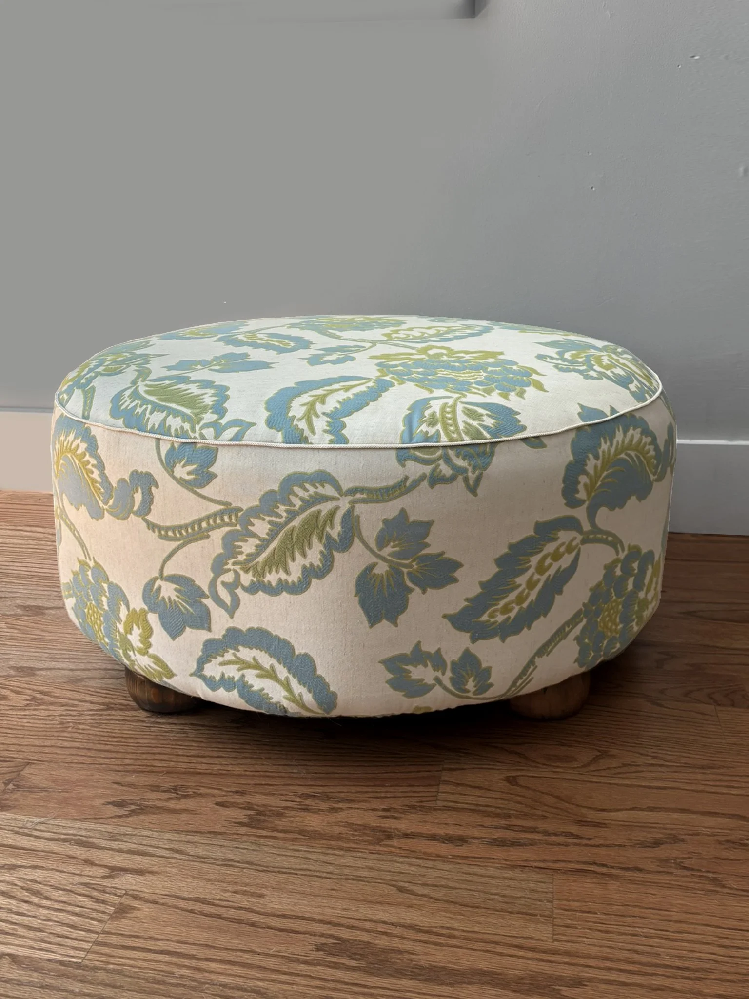 Round upholstered floral ottoman with blue and green botanical fabric on wooden legs
