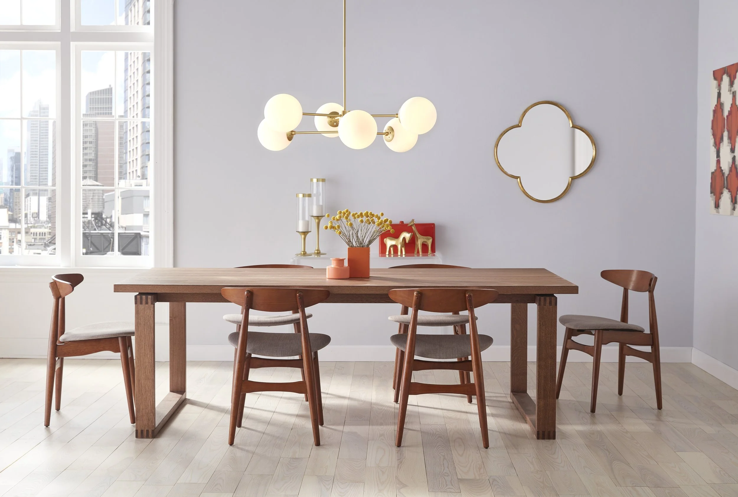 Bright mid-century modern dining room with a wooden table, four chairs, globe pendant light, decorative wall mirror, and city views through large windows