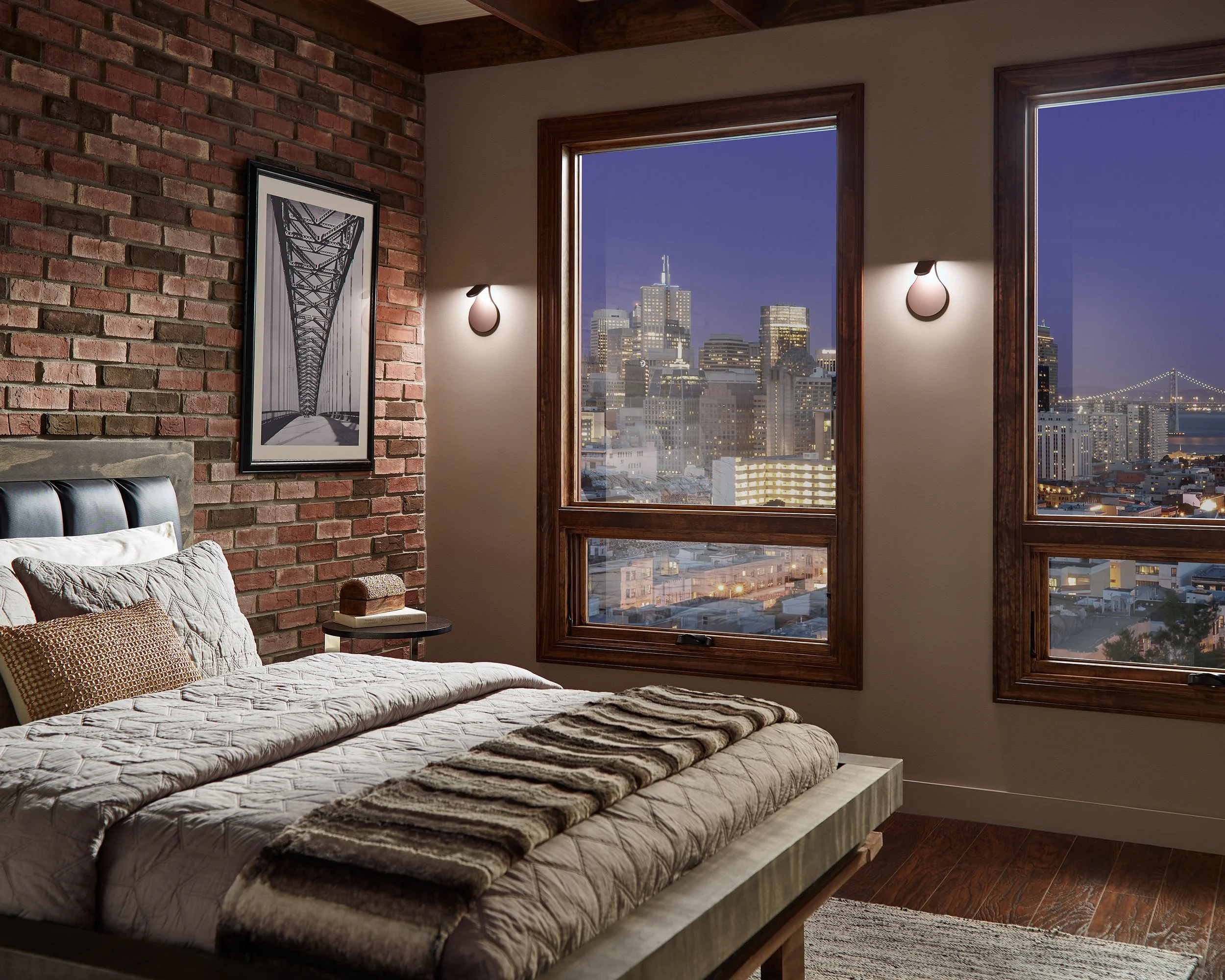 Modern bedroom with brick accent wall, large city-view windows, platform bed, framed bridge photograph, and evening skyline