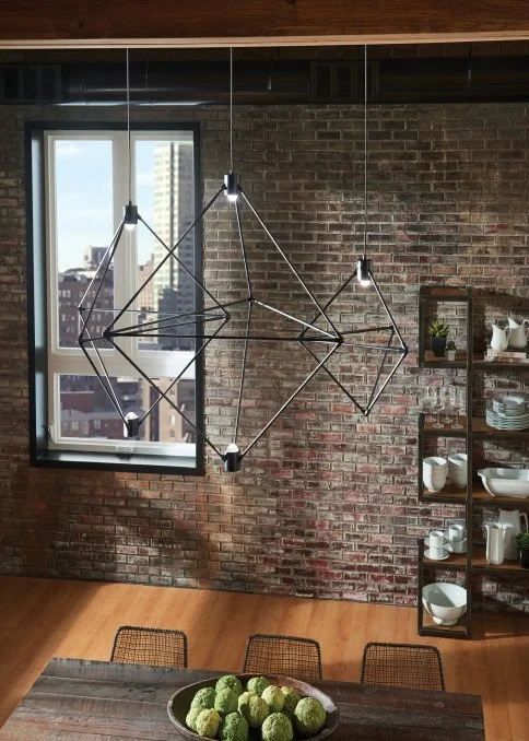 Modern geometric black pendant light fixture above a dining table in a brick-walled loft with open shelving and city views