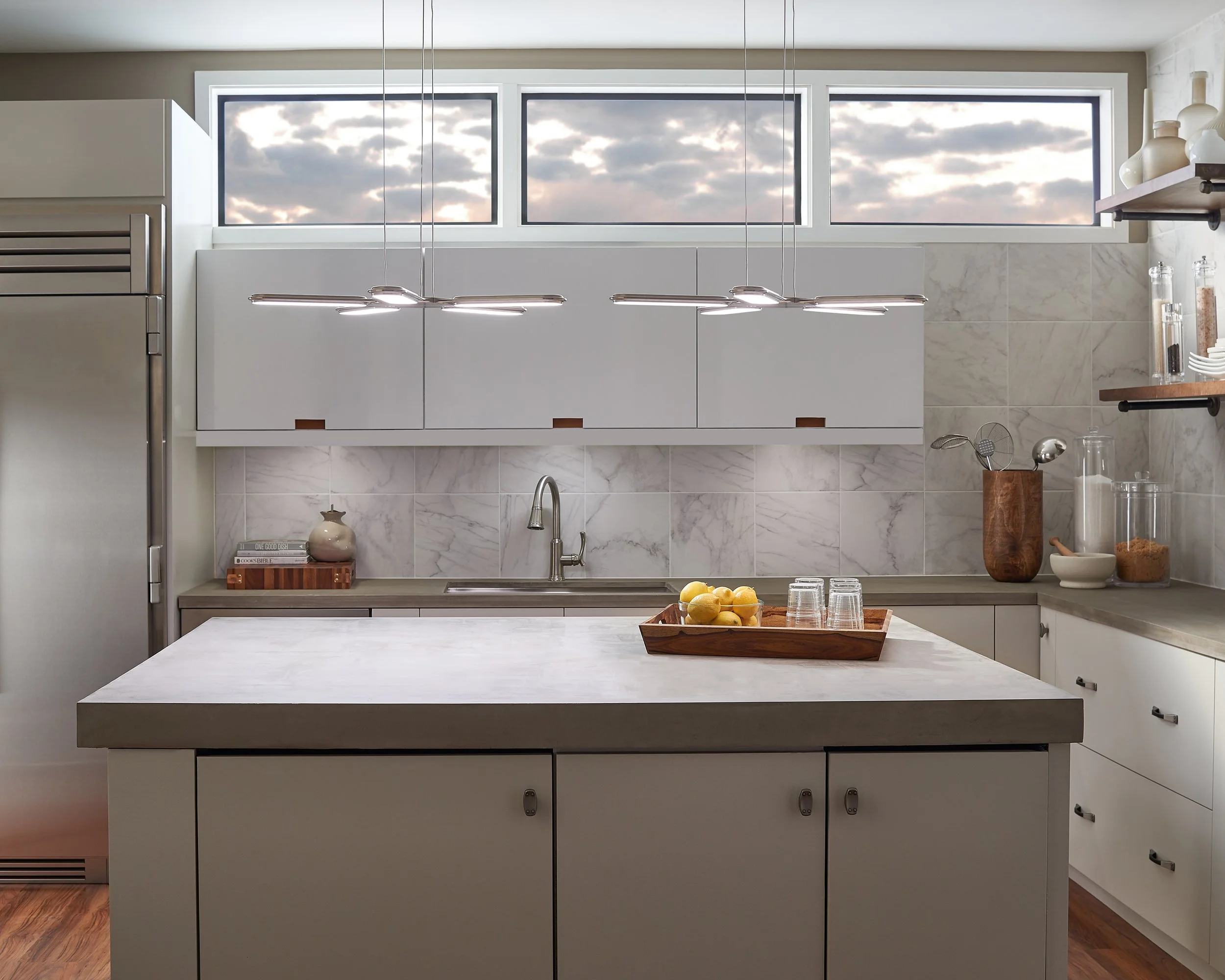 Modern kitchen with marble backsplash, central island, pendant lights, stainless steel refrigerator, sink, open shelves, and a bowl of lemons.