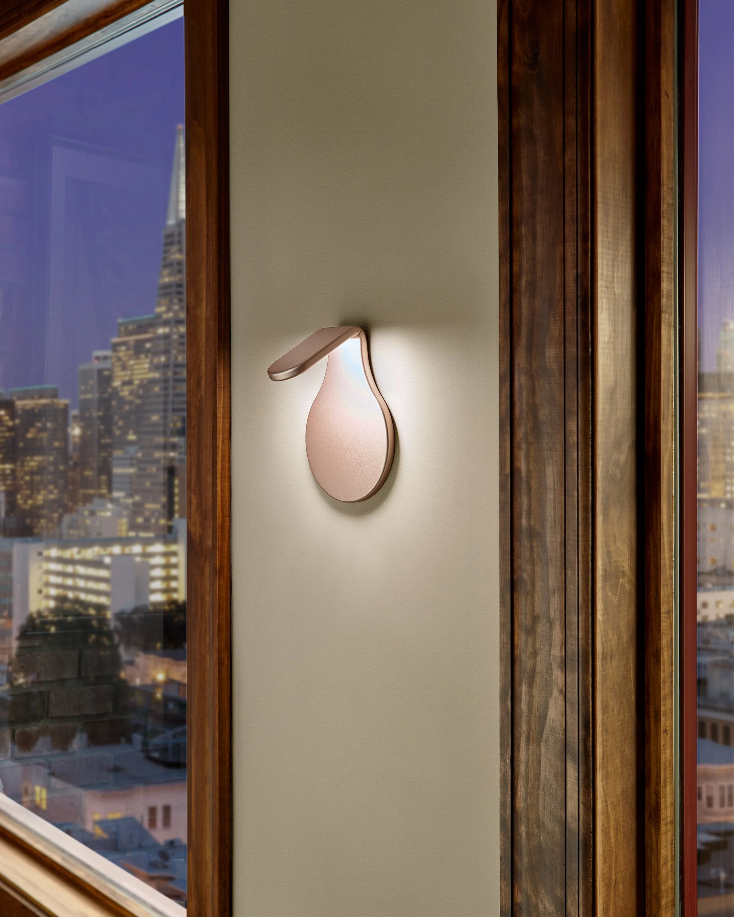 Modern wall sconce mounted on a textured wall between wood-framed windows overlooking a city skyline at night