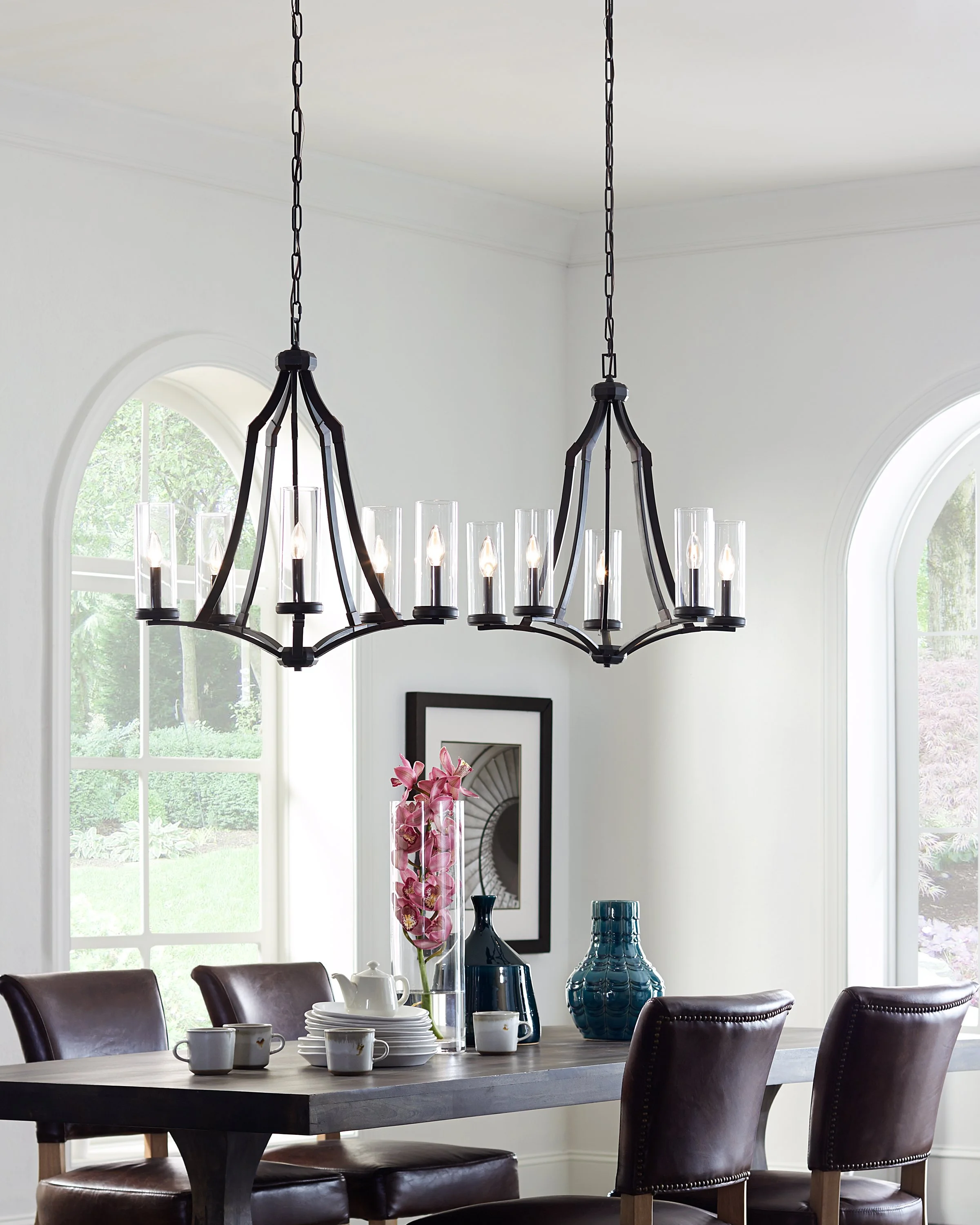 Elegant dining room with a black multi-light chandelier, dark leather chairs, tableware, blue vases, and arched windows