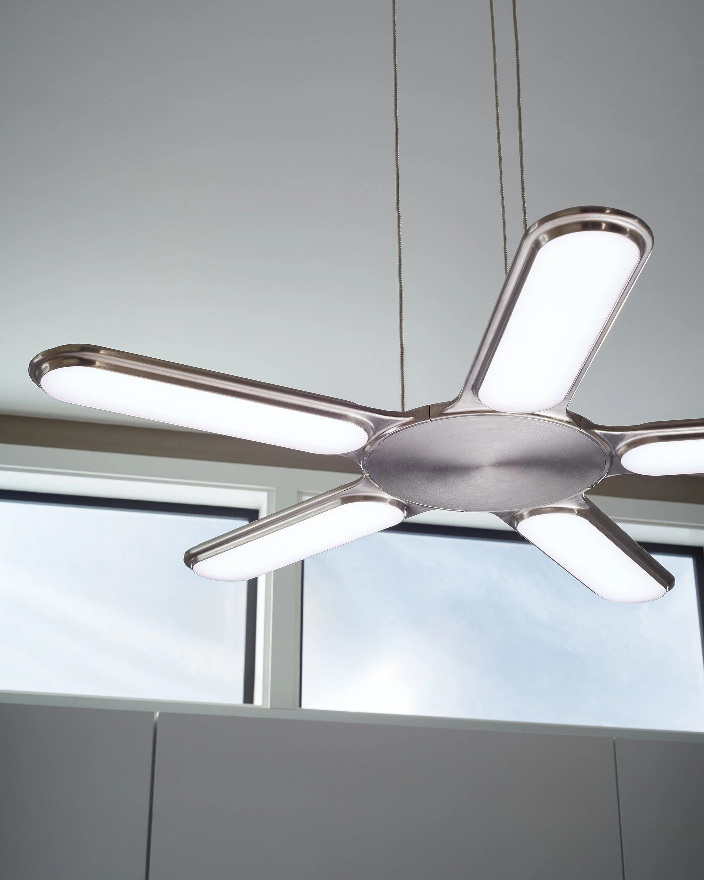 Modern brushed-nickel ceiling fan with five illuminated blades hanging in front of large windows