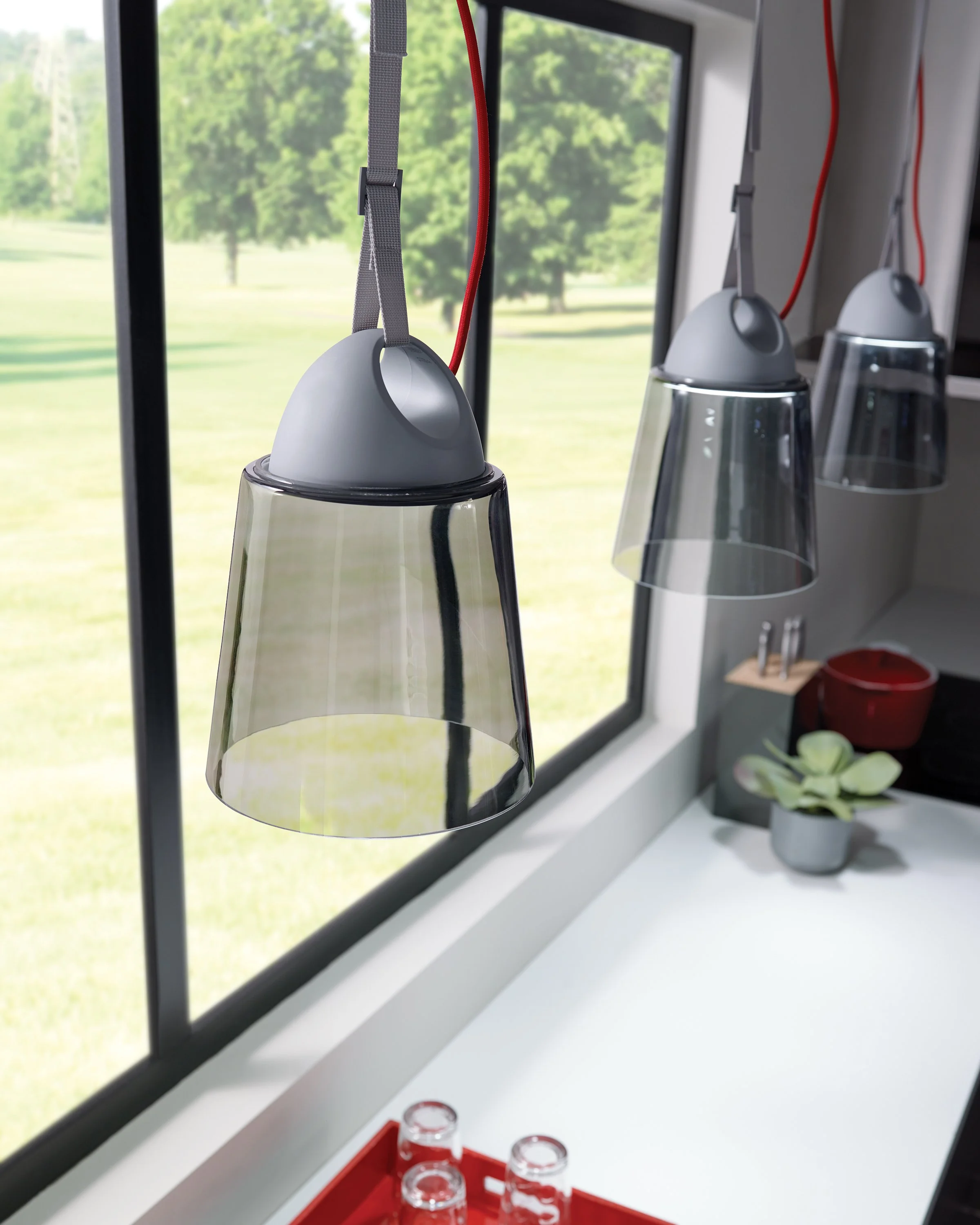 Modern hanging pendant lights beside a large window overlooking a green lawn, with a potted plant and red tray on the sill