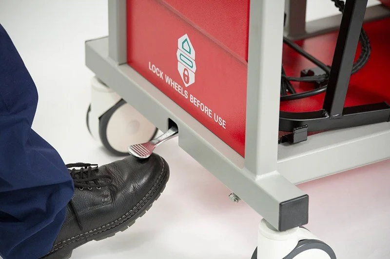 Foot-operated wheel lock pedal on a red medical equipment cart labeled “Lock Wheels Before Use.”
