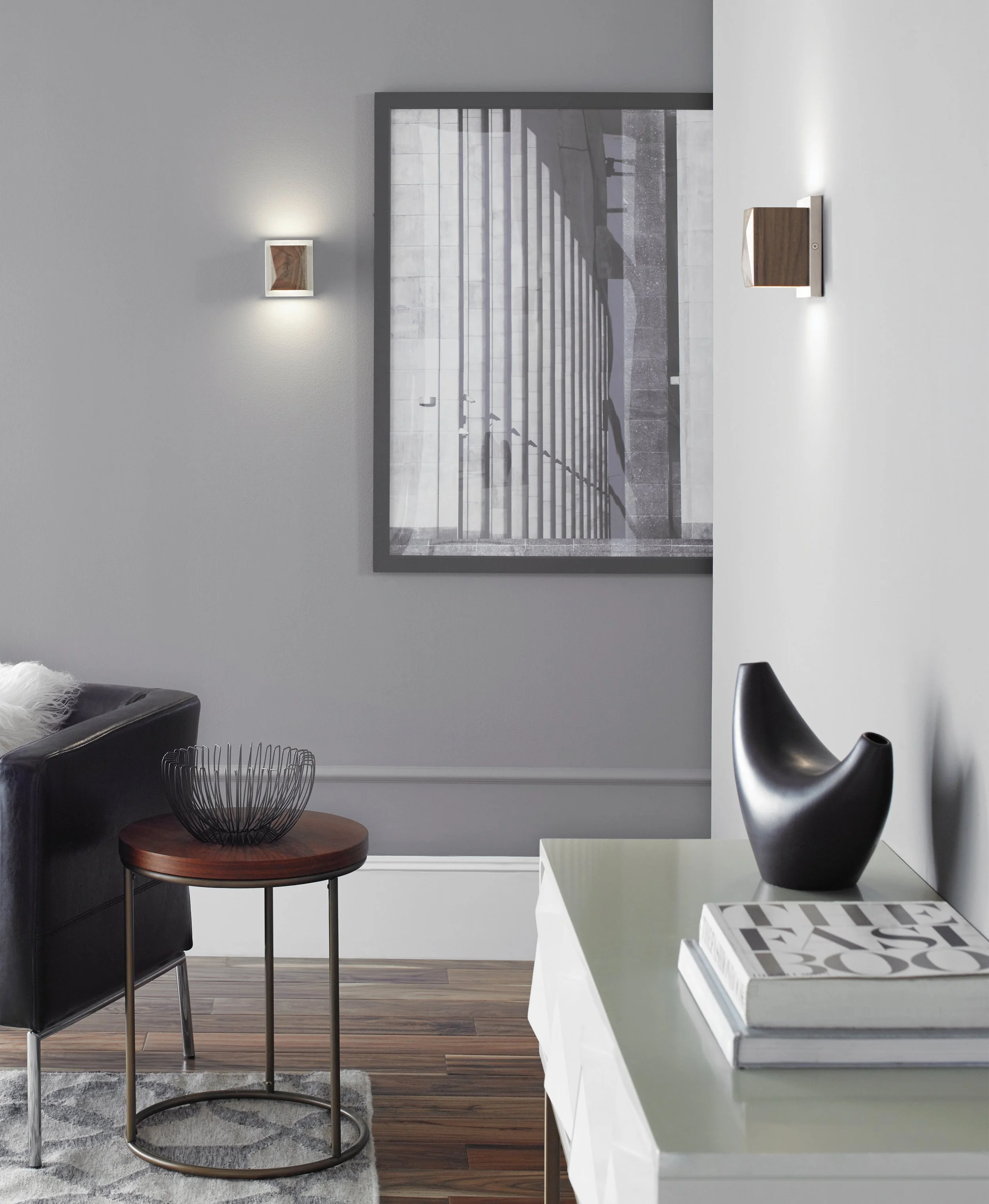 Modern living room with framed architectural artwork, black leather chair, round side table, wire basket, and sculptural black vase