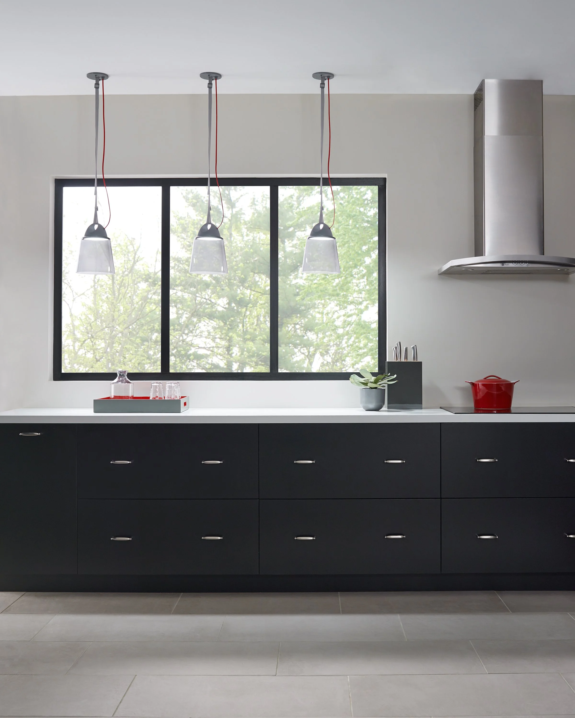 Modern black-and-white kitchen with three pendant lights, large window, stainless steel range hood, red Dutch oven, and black cabinets.