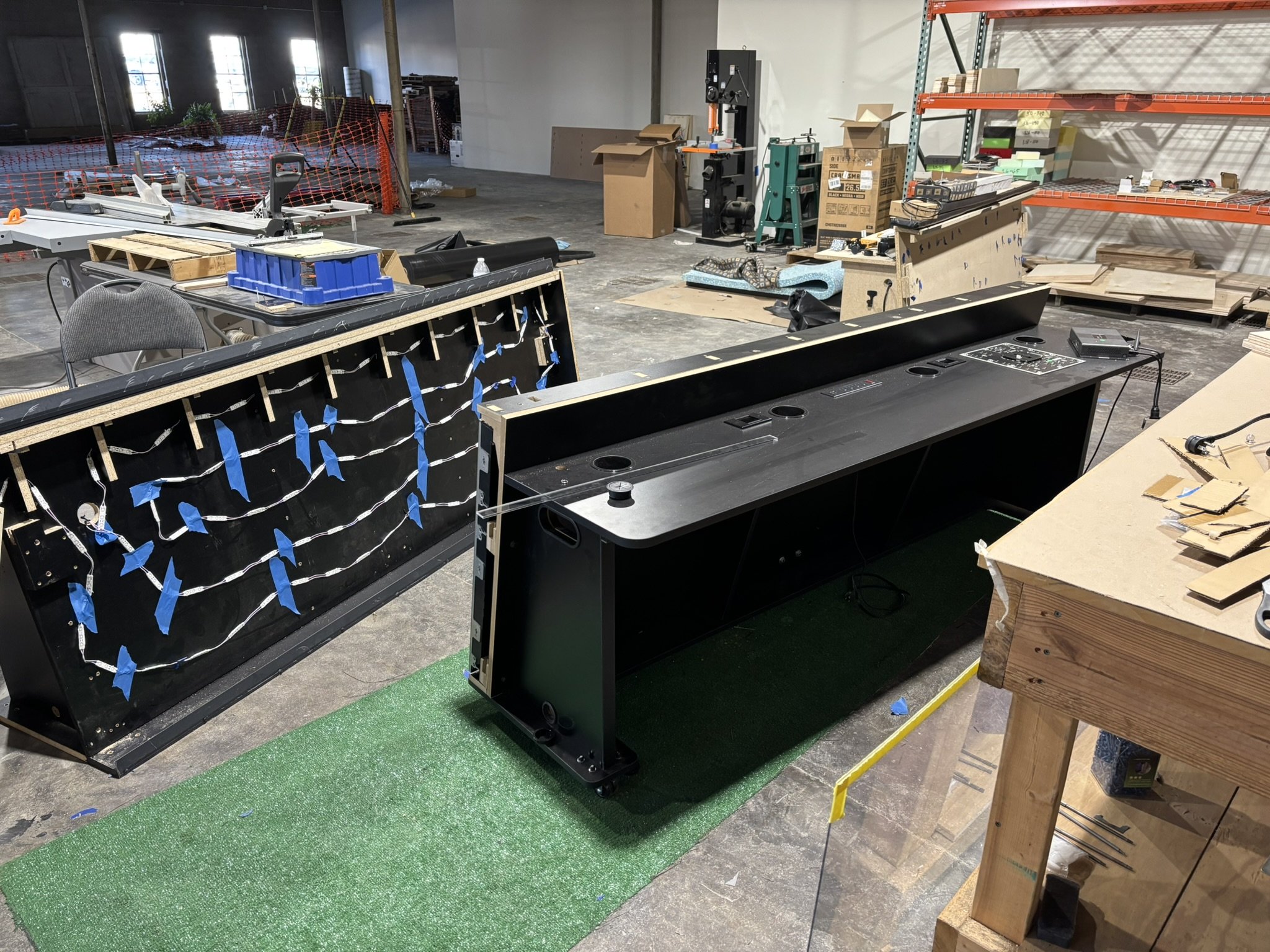 Workshop filled with partially assembled black arcade cabinets, LED wiring, tools, cardboard boxes, and industrial storage shelving.