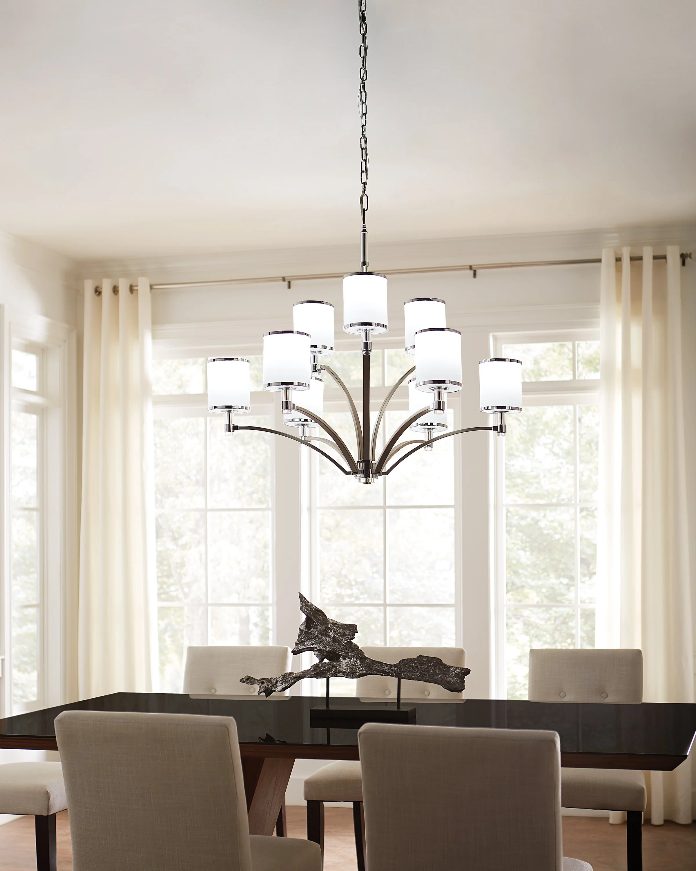 Modern dining room with a multi-light chrome chandelier, white upholstered chairs, large windows, and a sculptural centerpiece