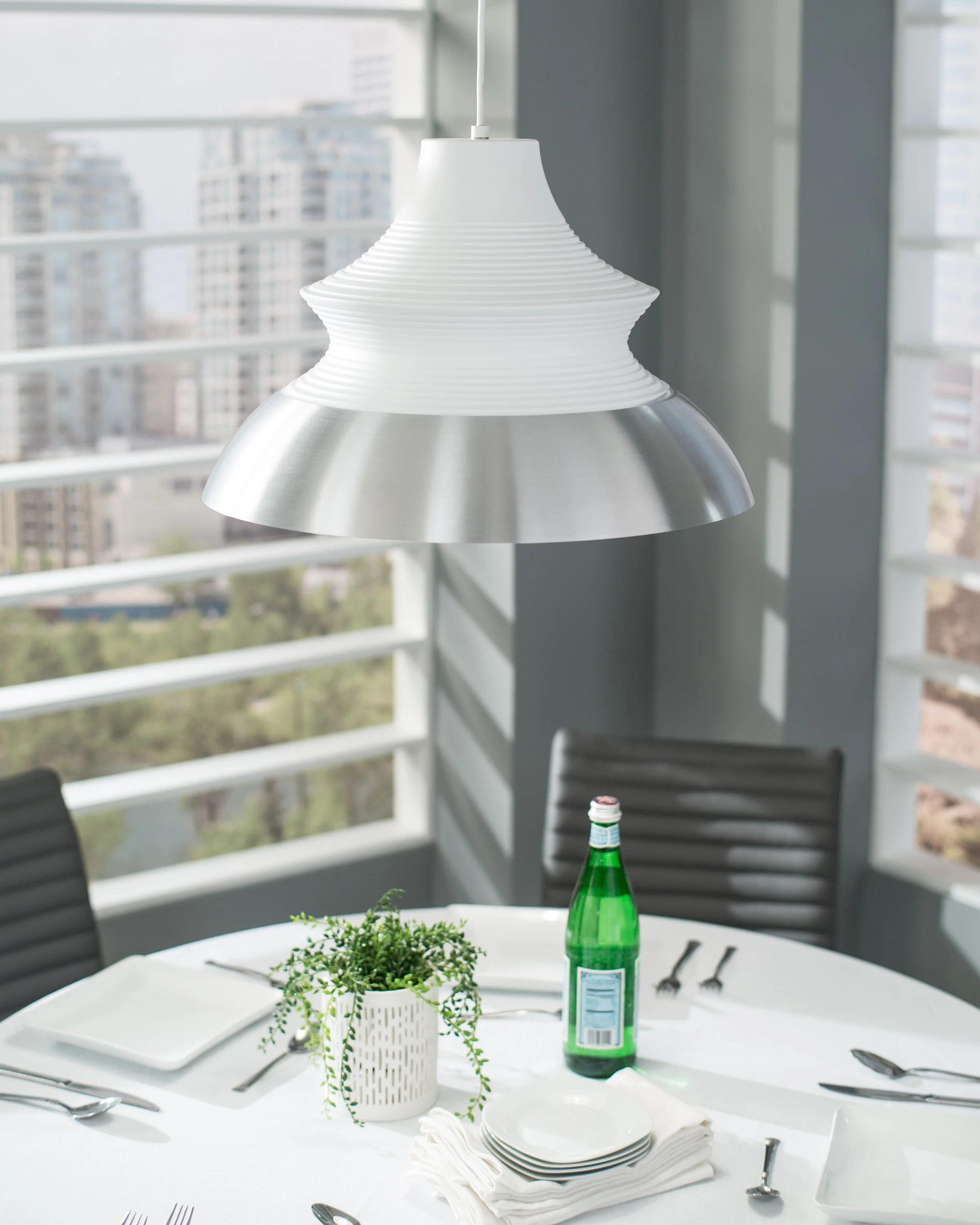 White and silver pendant light hanging over a set dining table with a potted plant and green bottle by large windows