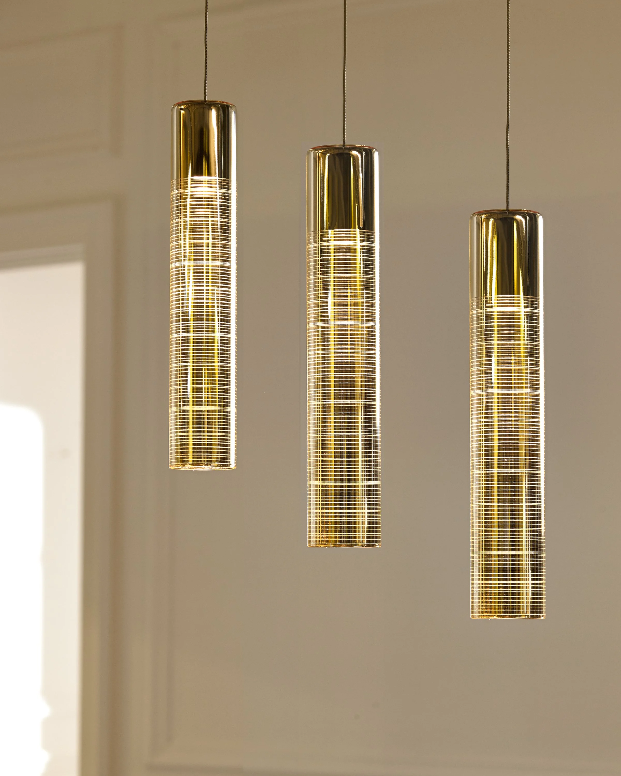 Three illuminated gold cylindrical pendant lights hanging at different heights against a beige wall.