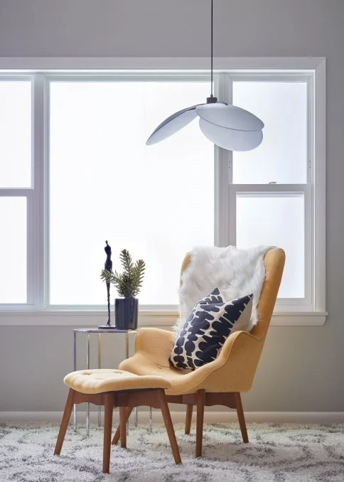 Midcentury modern mustard-yellow armchair with white sheepskin throw and patterned pillow beside a matching ottoman, pendant lamp, and plant by a bright window