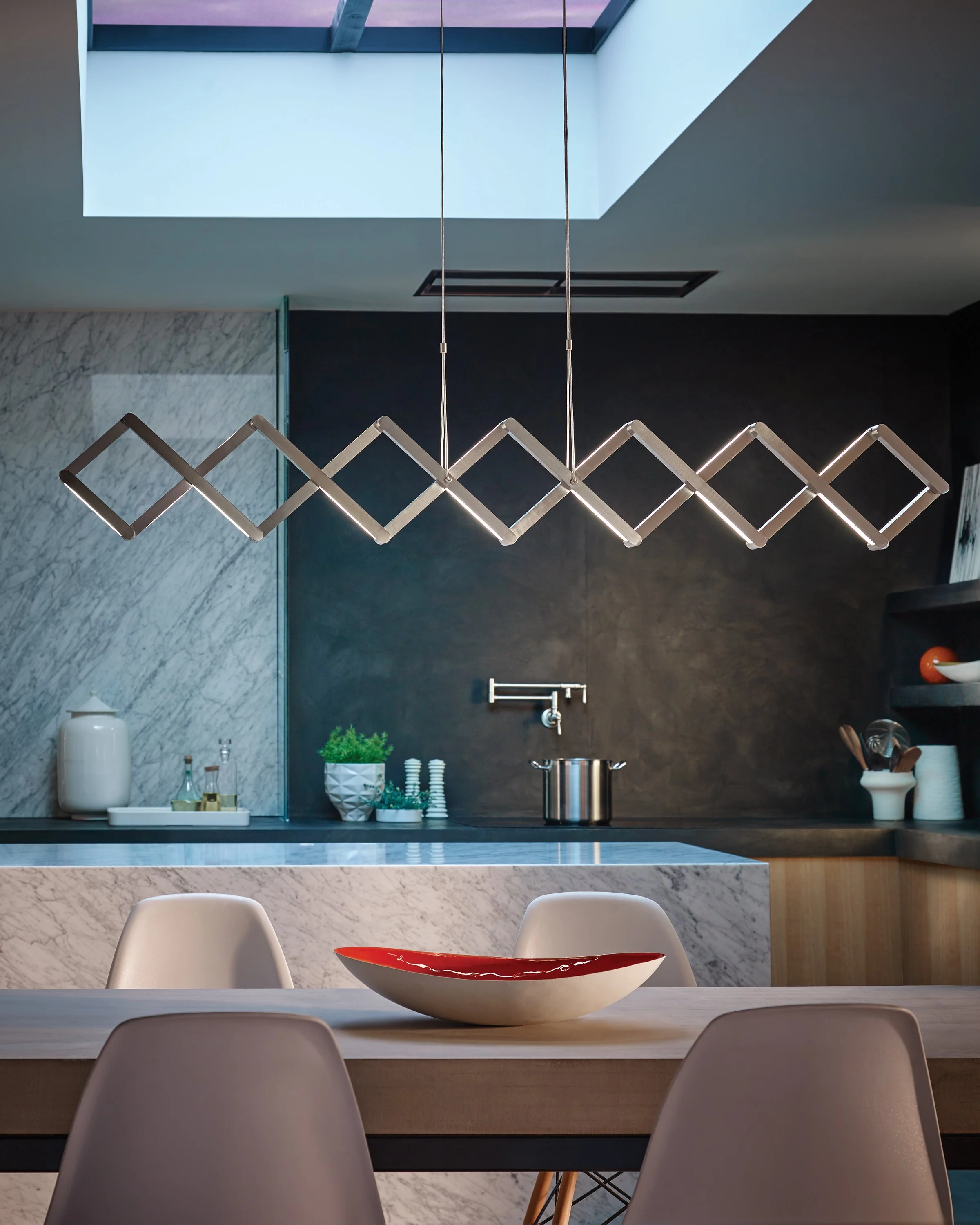 Modern kitchen and dining area with marble island, geometric pendant light, dark cabinetry, bar stools, and decorative bowl