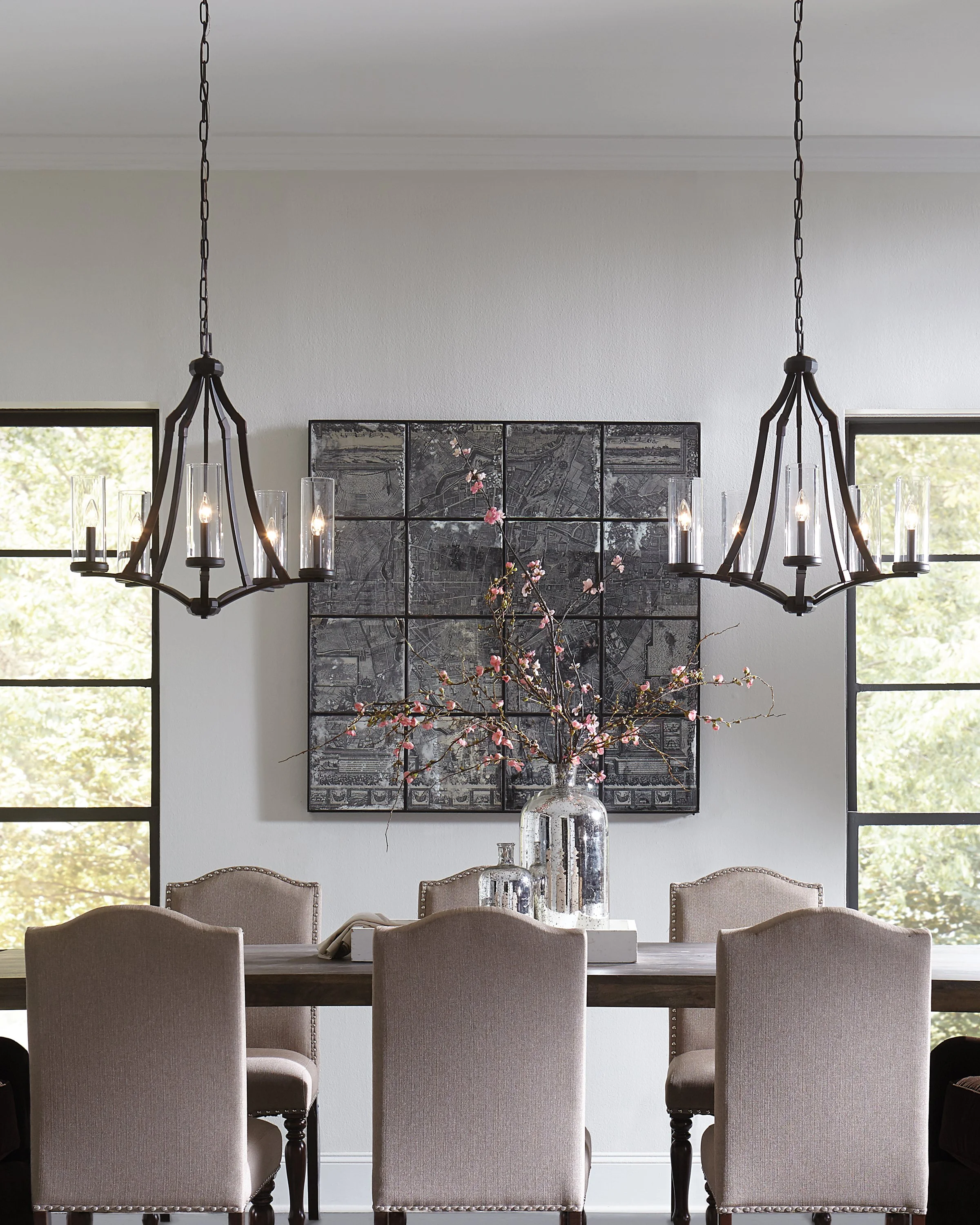 Elegant dining room with two black chandeliers, a large vintage map artwork, flowering branches, and upholstered dining chairs.