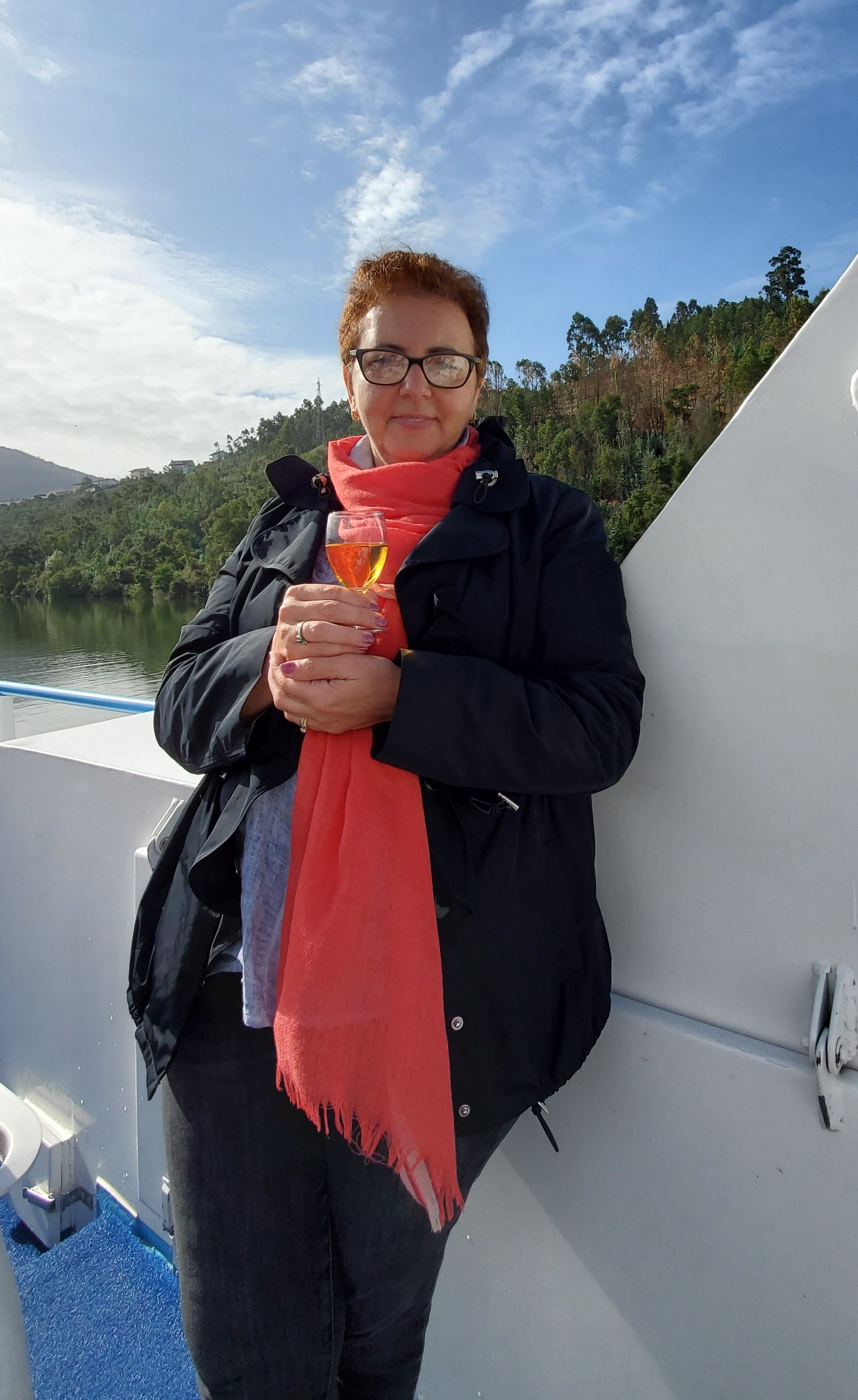 A woman with short reddish hair and glasses holding a glass of rosé wine on a boat. She is wearing a black jacket and a red scarf. In the background, there is a river with green hills and trees under a partly cloudy sky.