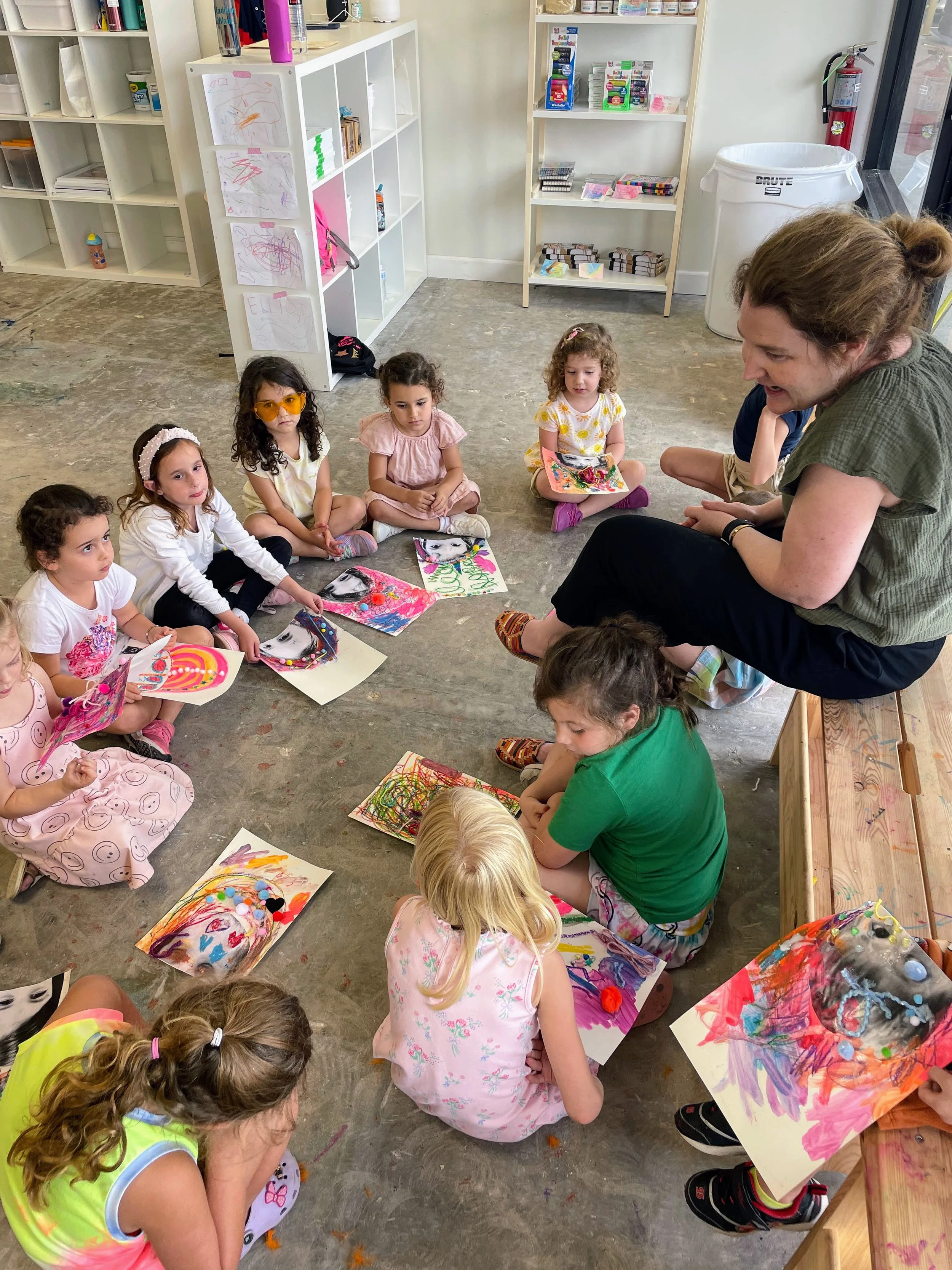 A group of young children sitting on the floor in a classroom circle, showing their colorful artwork, while an adult, likely a teacher, engages with them.