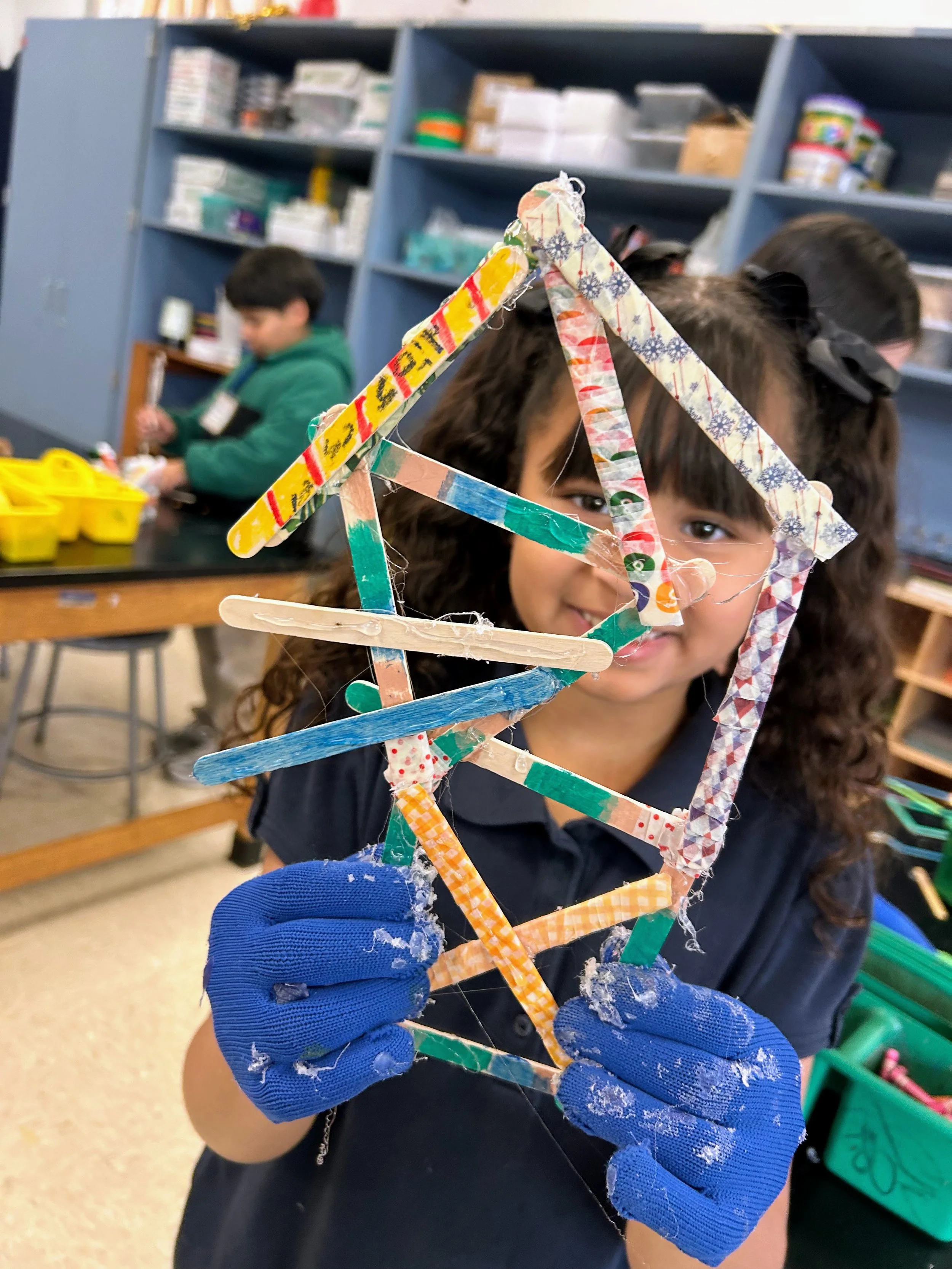 A young girl with curly brown hair and wearing blue gloves is holding up a colorful craft made of cut and decorated popsicle sticks. The craft appears to form a house or tree, and she is smiling while showing it to the camera. In the background, there are shelves with various craft supplies and other children working on projects.
