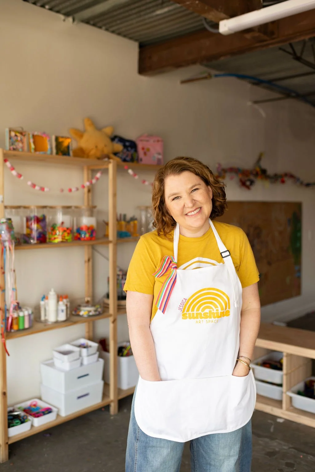 A smiling woman with curly hair wearing a yellow shirt and a white apron in an art studio or craft room.