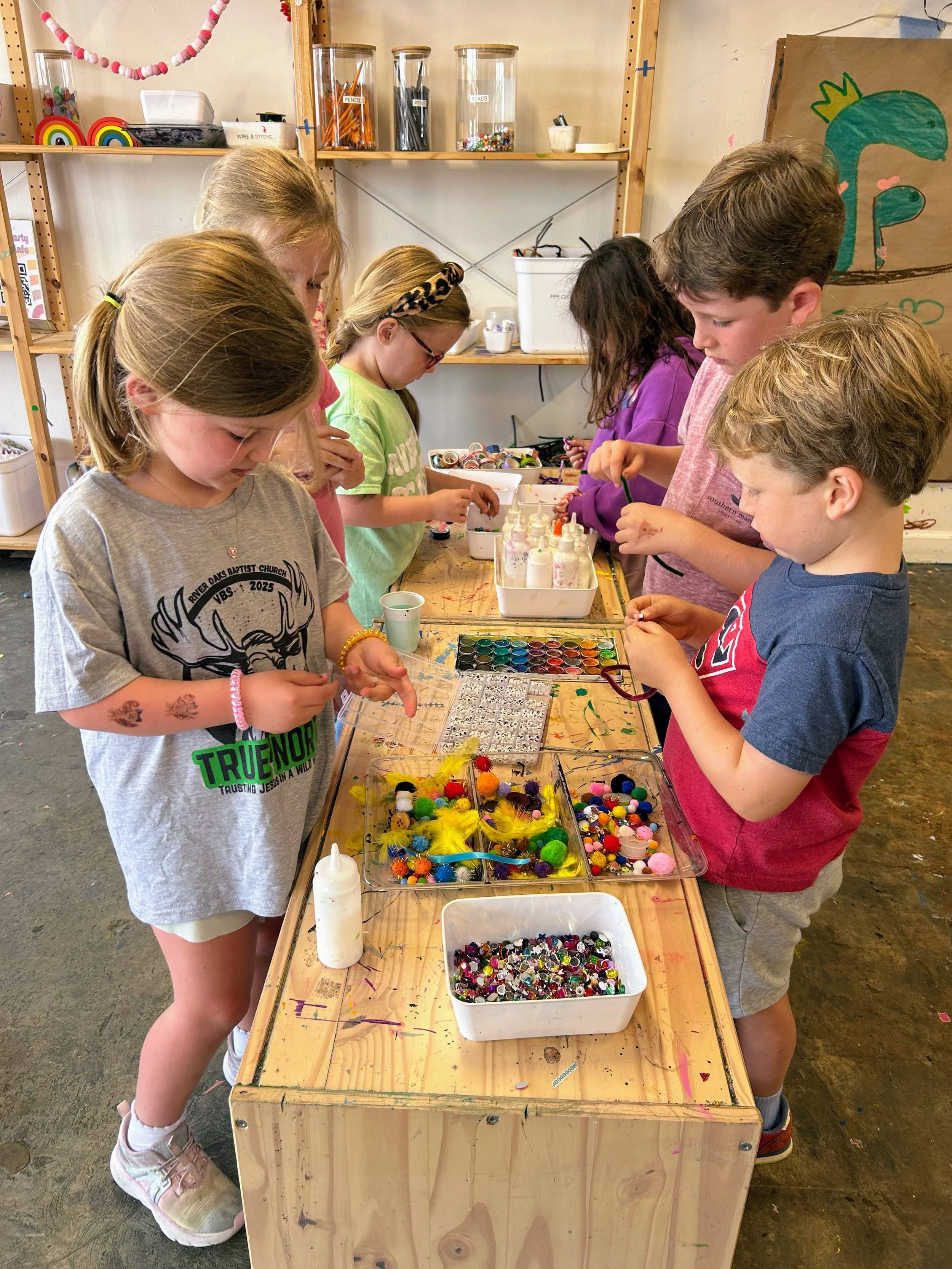 Group of children gathered around a wooden table with various colorful craft supplies, including pom-poms, rhinestones, and bottles of glue, in an art classroom.