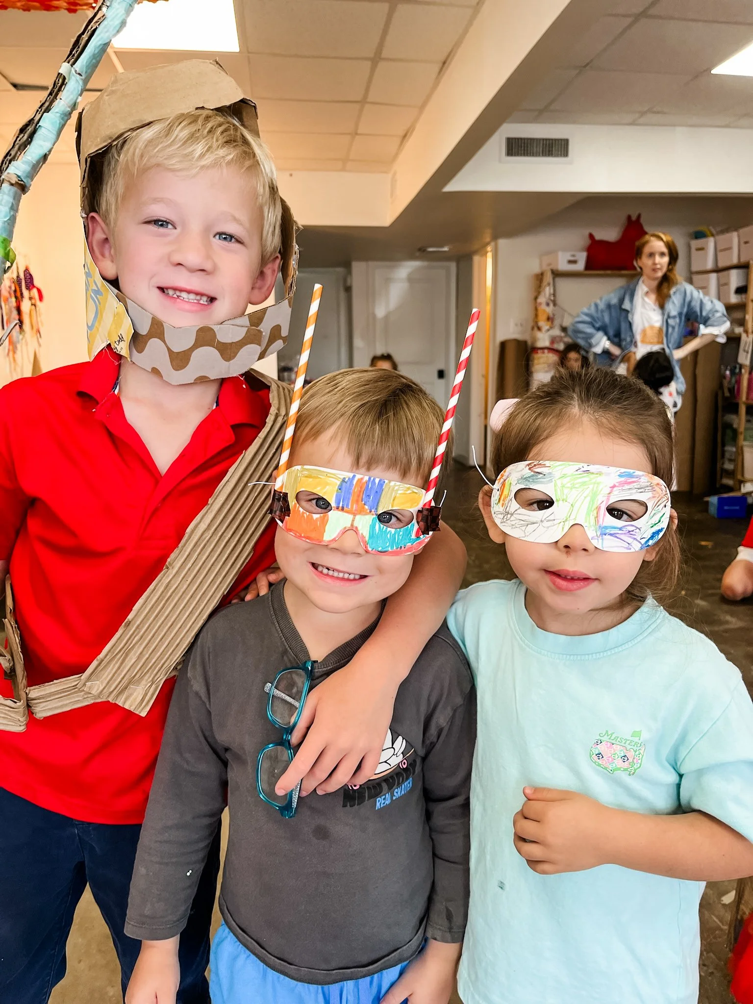 Four children wearing colorful masks and decorations for a costume event in an indoor setting, with a woman in background watching.