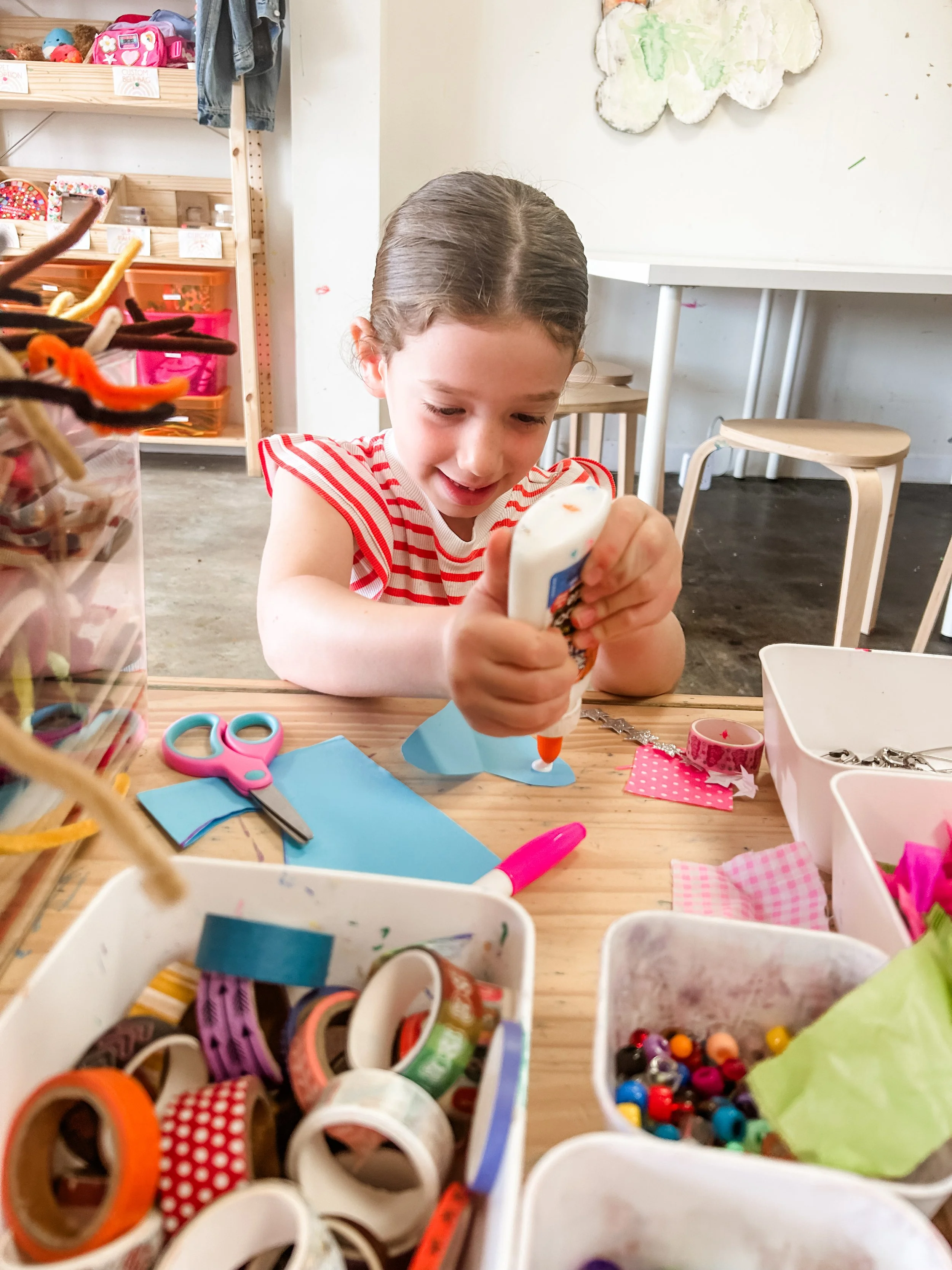 A young girl with brown hair in a ponytail sitting at a wooden table, smiling while squeezing glue onto a craft project. The table is cluttered with colorful crafting supplies including tape, scissors, paper, and beads. In the background, shelves with toys and a wall with a painted cloud.
