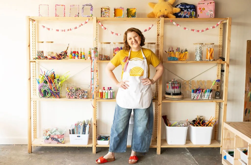 A woman standing in front of shelves filled with art supplies and crafts in a classroom or craft room.