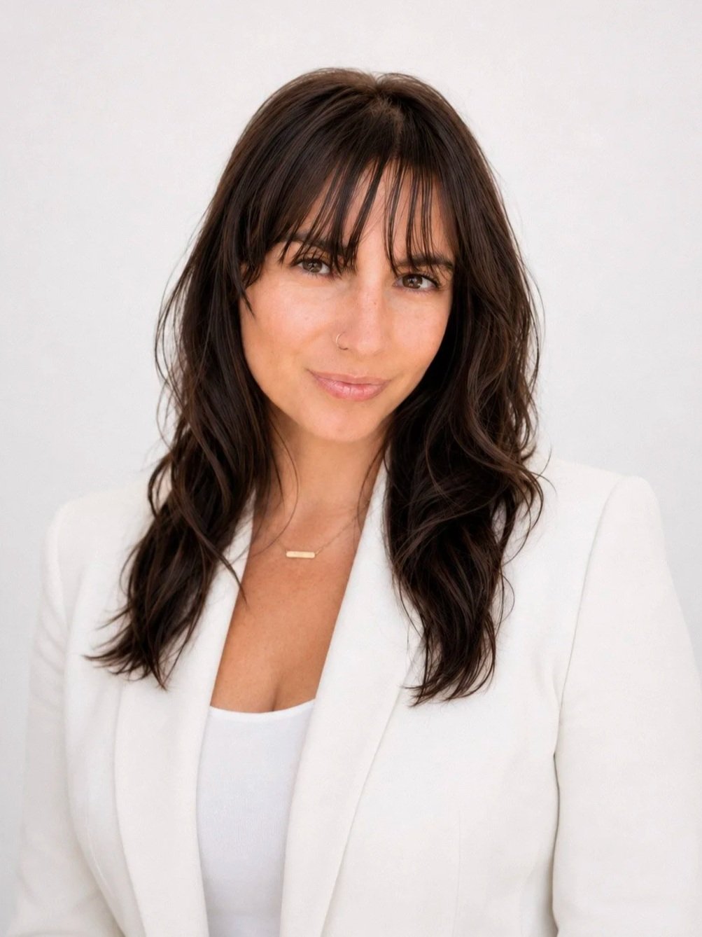 A woman with shoulder-length dark brown hair, wearing a white blazer and a white top, posing against a light-colored background.