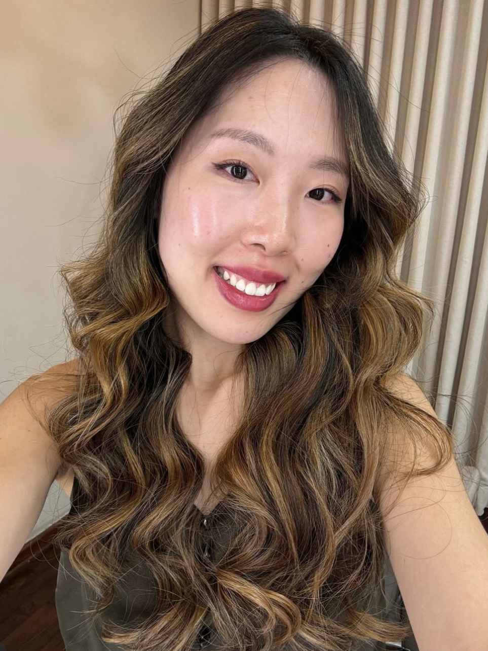 A smiling woman with long, wavy hair taking a selfie indoors.