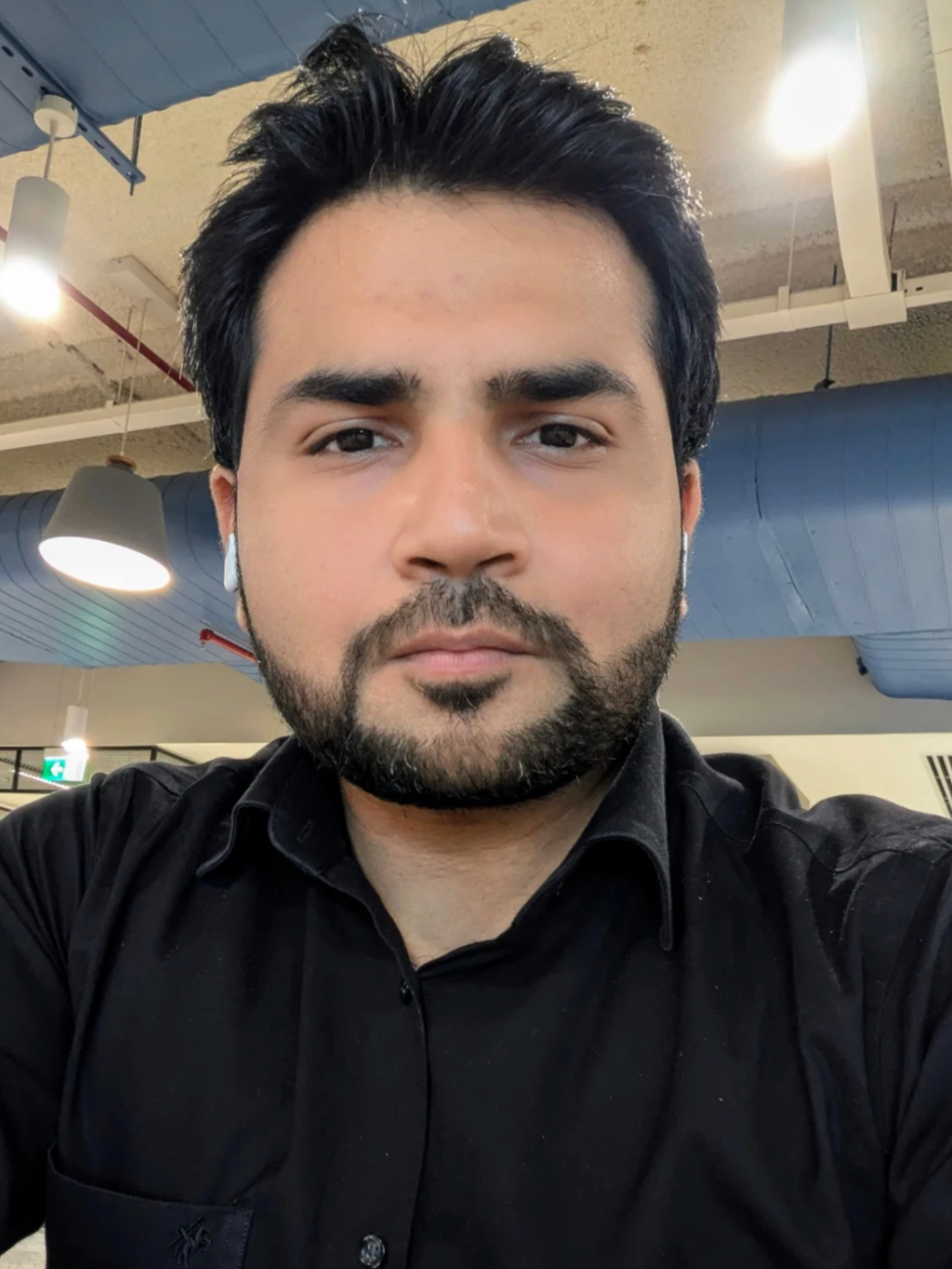 Close-up of a man with dark hair, beard, and mustache wearing a black shirt, indoors with ceiling lights and ventilation ducts visible.