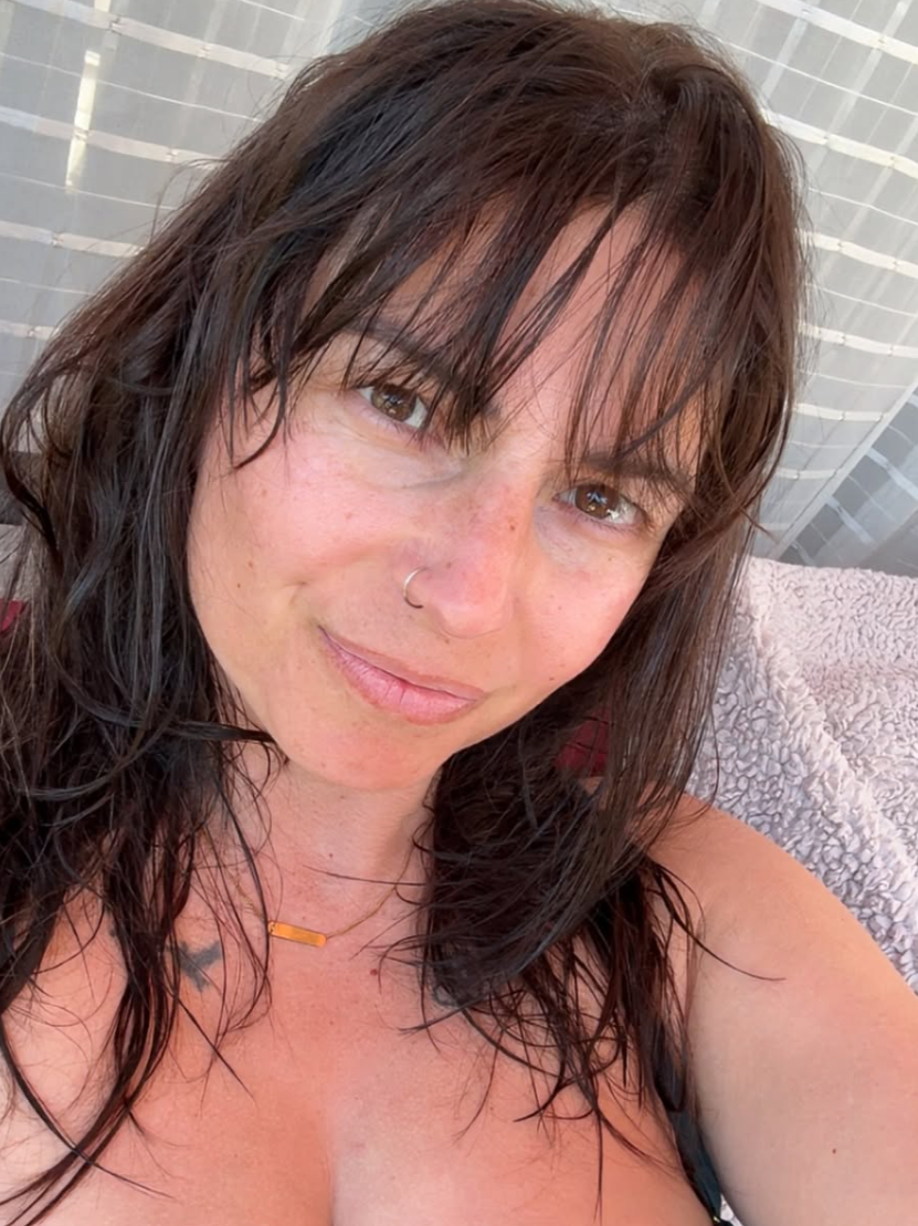 Close-up of a woman with brown, wavy hair, wearing a nose ring and a gold necklace, smiling slightly. She is outdoors, sitting on a blanket or towel, with sheer curtains in the background.