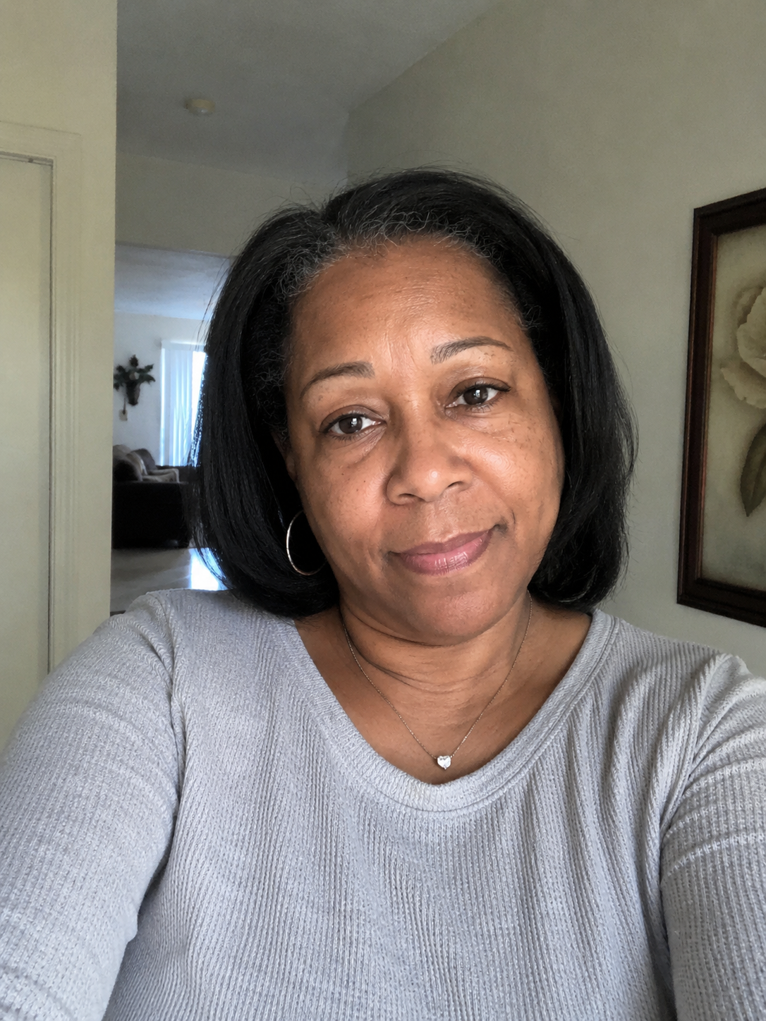 A woman with shoulder-length black hair wearing a light gray sweater and a heart-shaped pendant necklace, taking a selfie indoors with a living room and framed floral art in the background.
