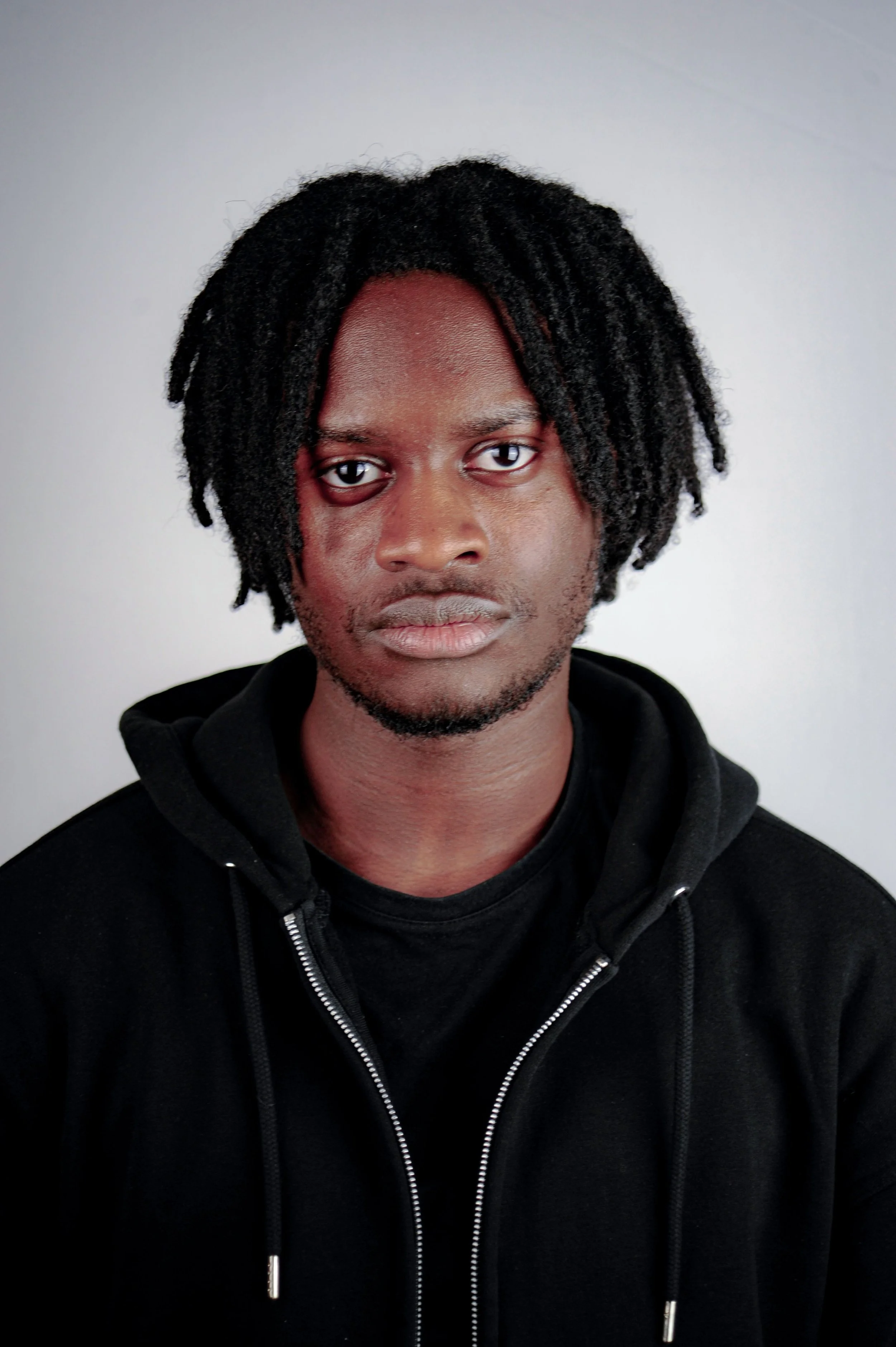 A young man with dreadlocks wearing a black hoodie and looking directly at the camera with a neutral expression against a plain light gray background.