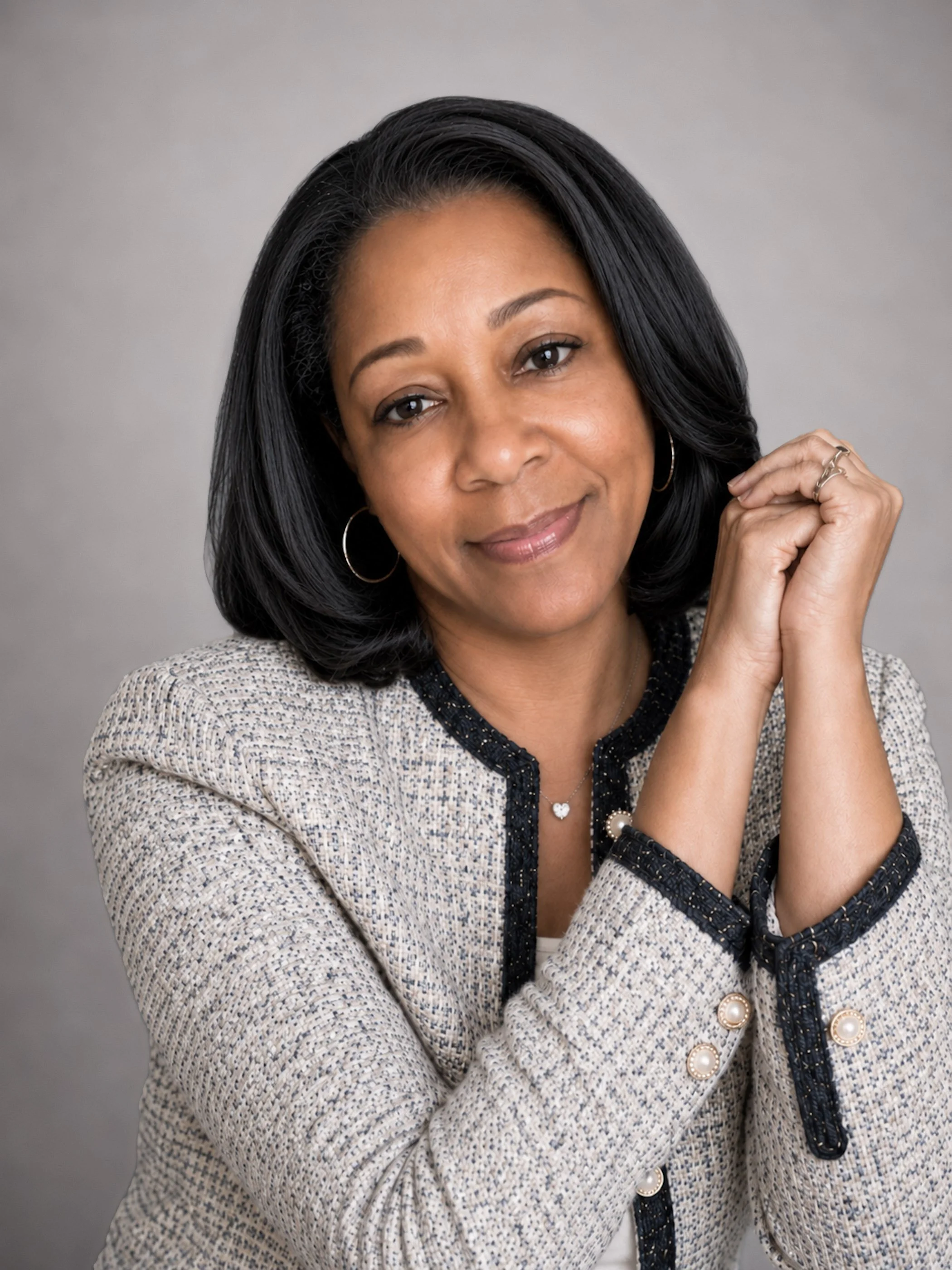 Close-up portrait of a smiling woman with black hair, wearing a patterned jacket with pearl buttons, earrings, necklace, and rings, against a gray background.